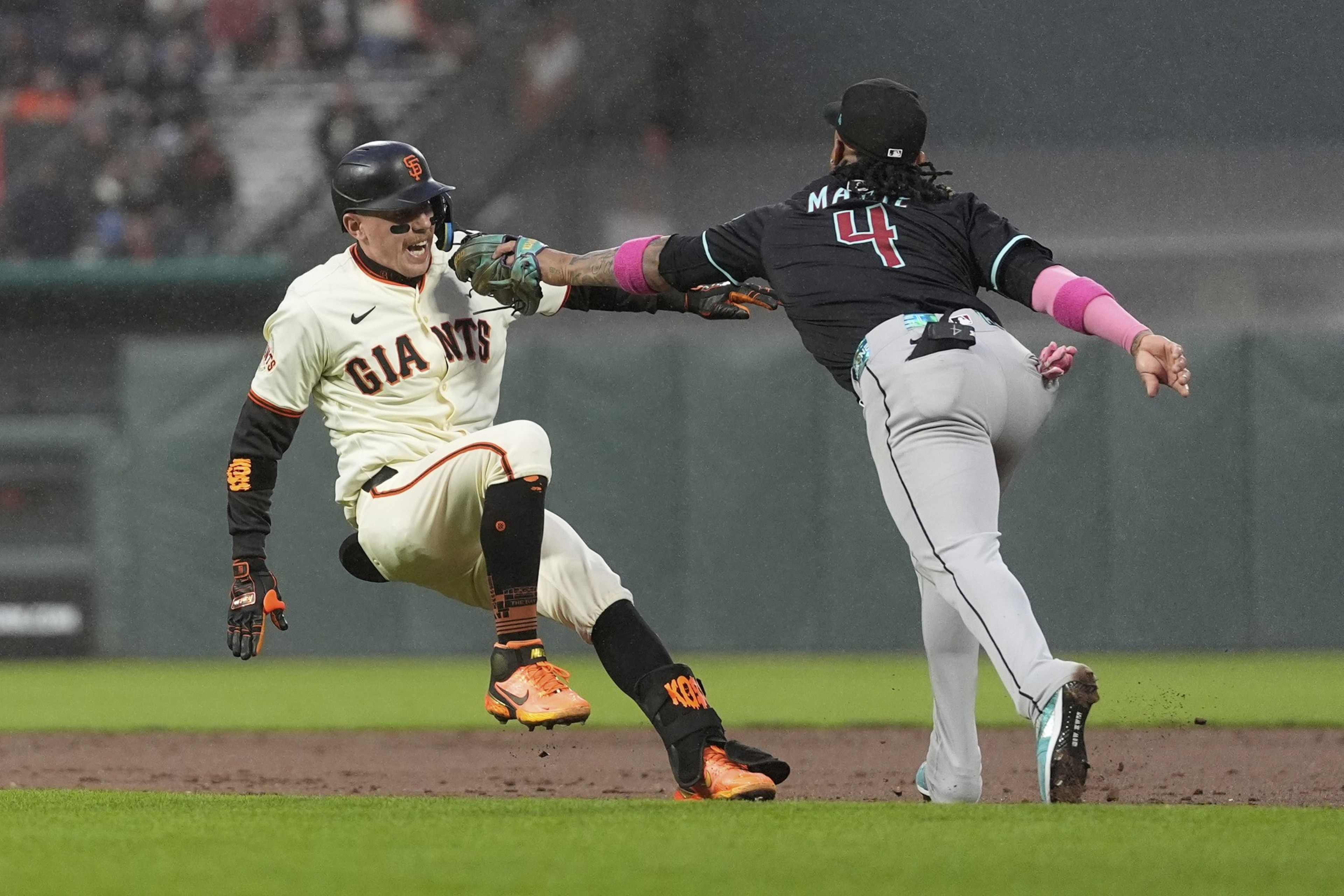 A baseball player in a white "Giants" uniform slides as an opposing player in a black and gray uniform tags him. The moment is action-packed and dynamic.