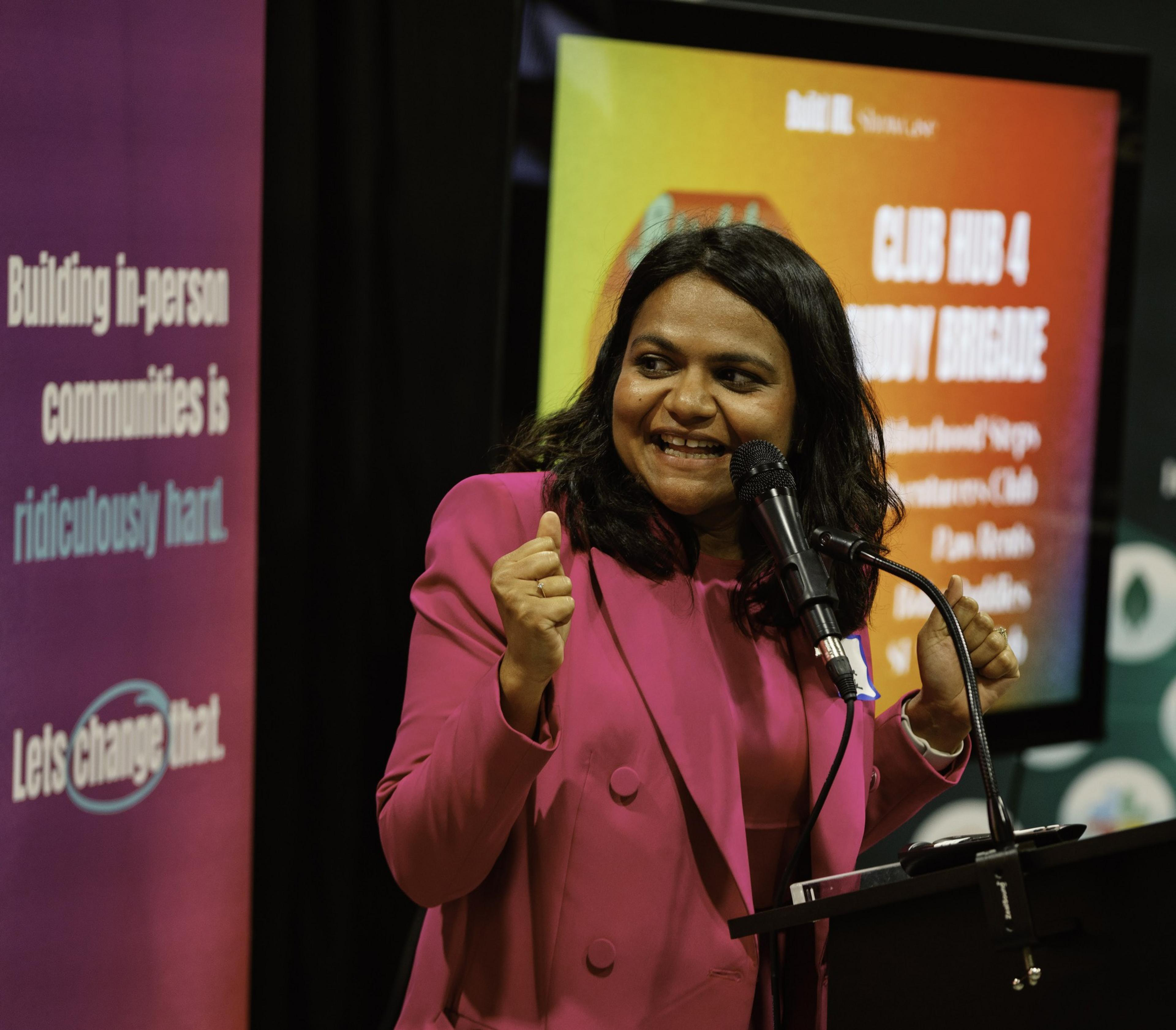 A woman in a bright pink suit is speaking enthusiastically at a podium with a microphone. A colorful poster and sign in the background contain text.