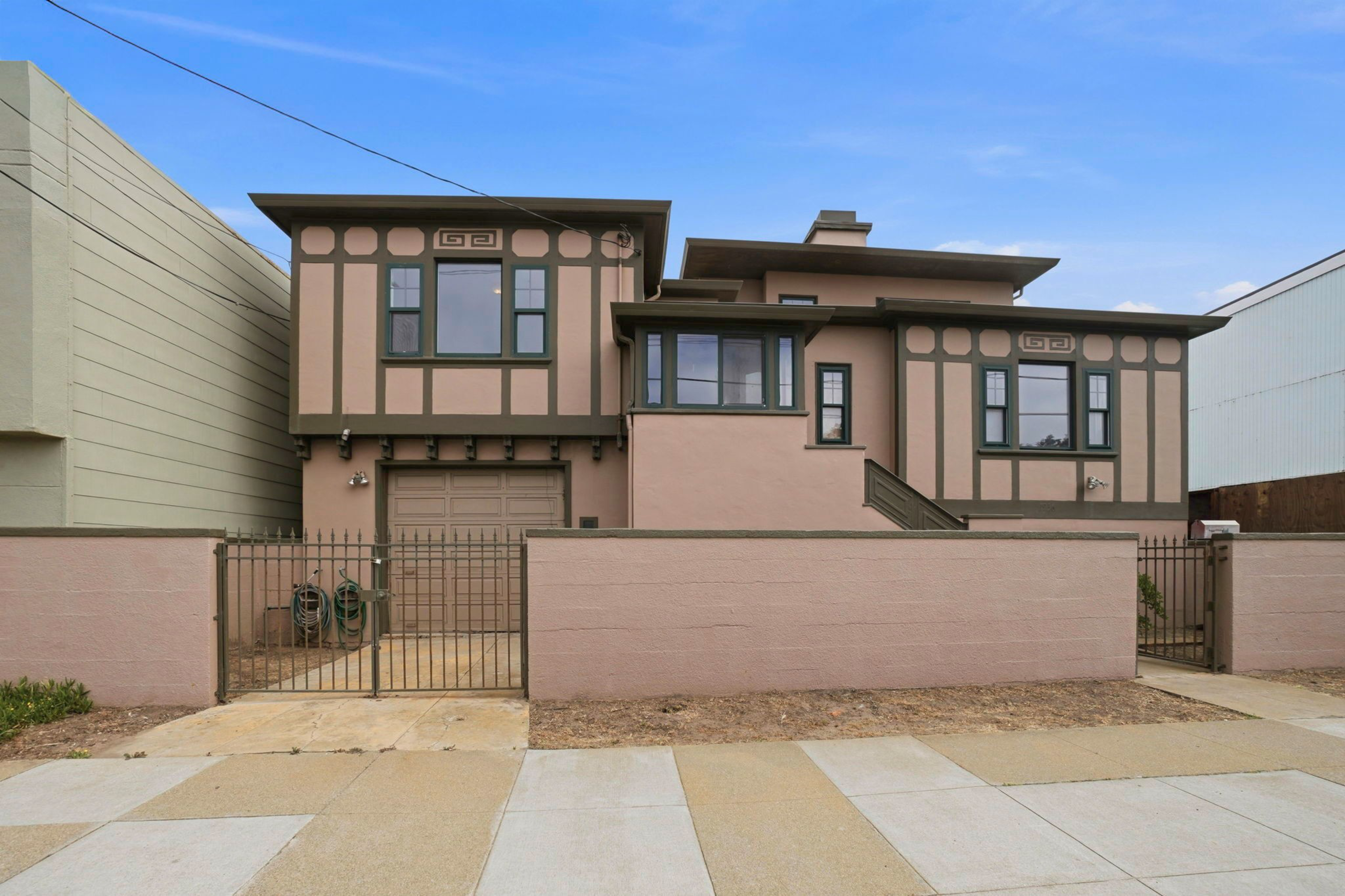 A two-story beige house with dark brown trim features multiple windows, a gated front yard, a garage, and minimal landscaping under a clear blue sky.
