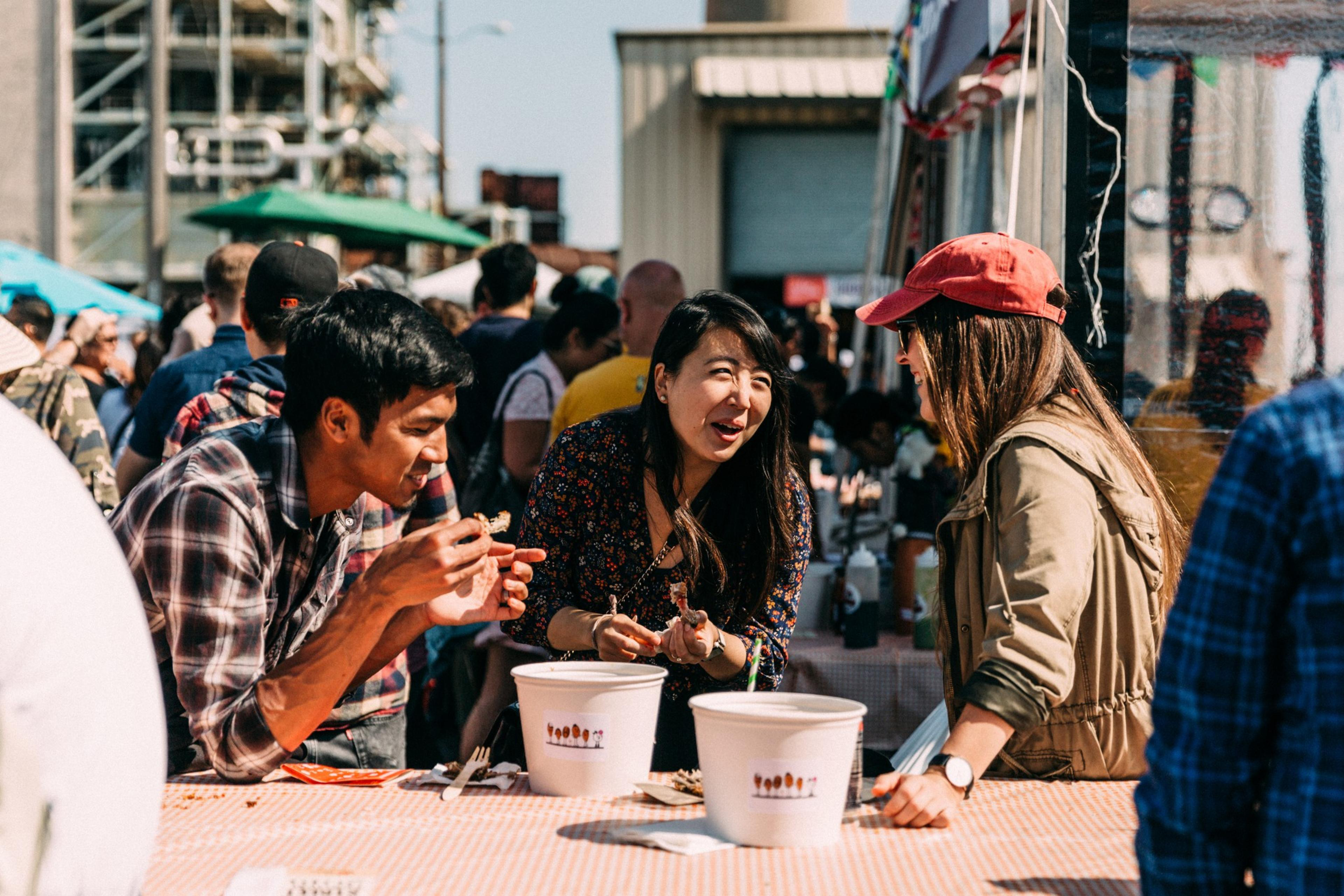 Three people are eating and talking animatedly at an outdoor food stall crowded with people on a sunny day.