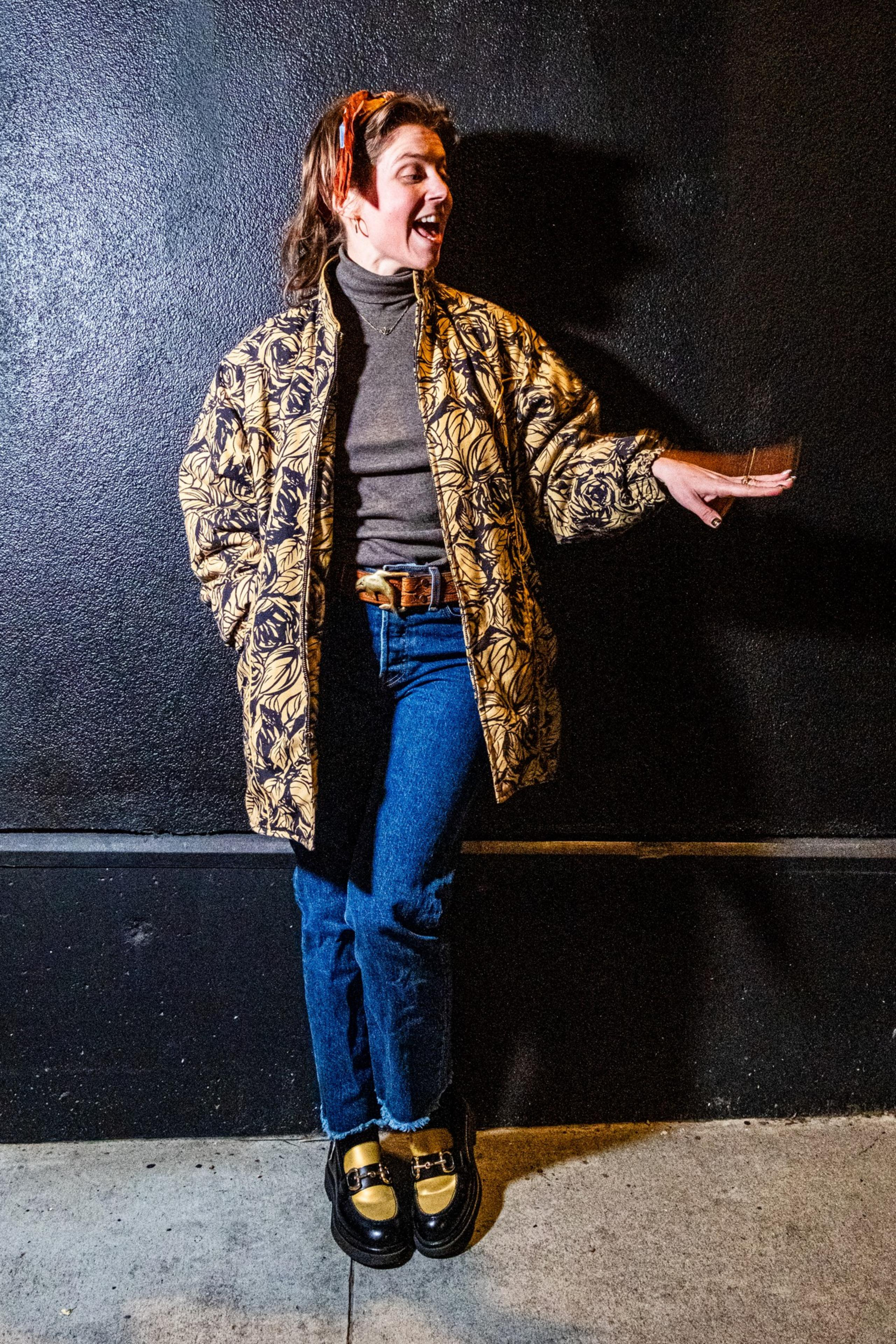 A woman stands against a textured black wall, wearing a patterned jacket, gray turtleneck, jeans, and loafers, with a joyful expression and hand extended.