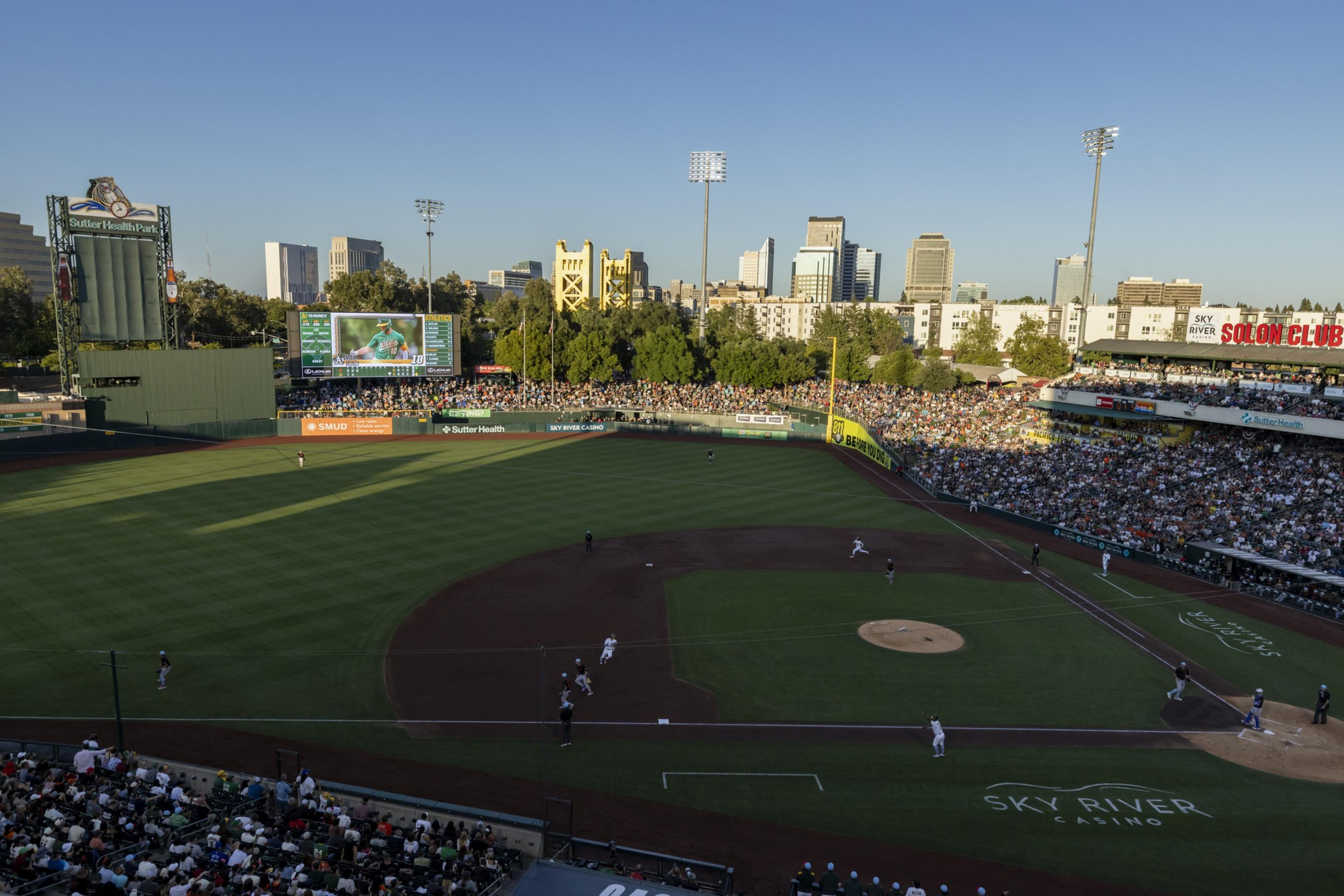 A baseball game is being played at a stadium with a green field and packed stands. The skyline and a golden tower are visible in the background.