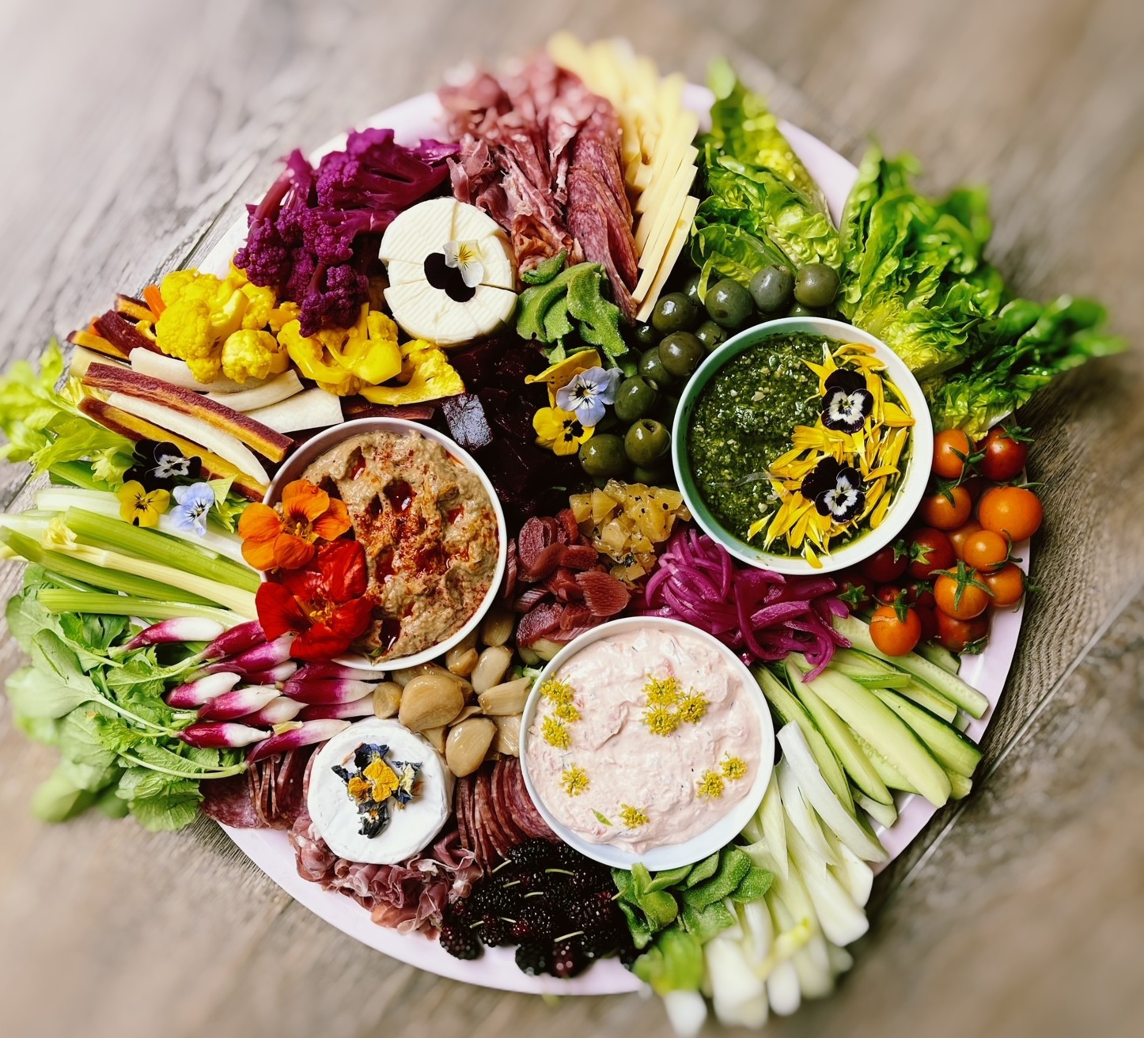 A colorful platter with various fresh vegetables, olives, three dips, cheeses, cured meats, and edible flowers arranged artfully on a wooden table.