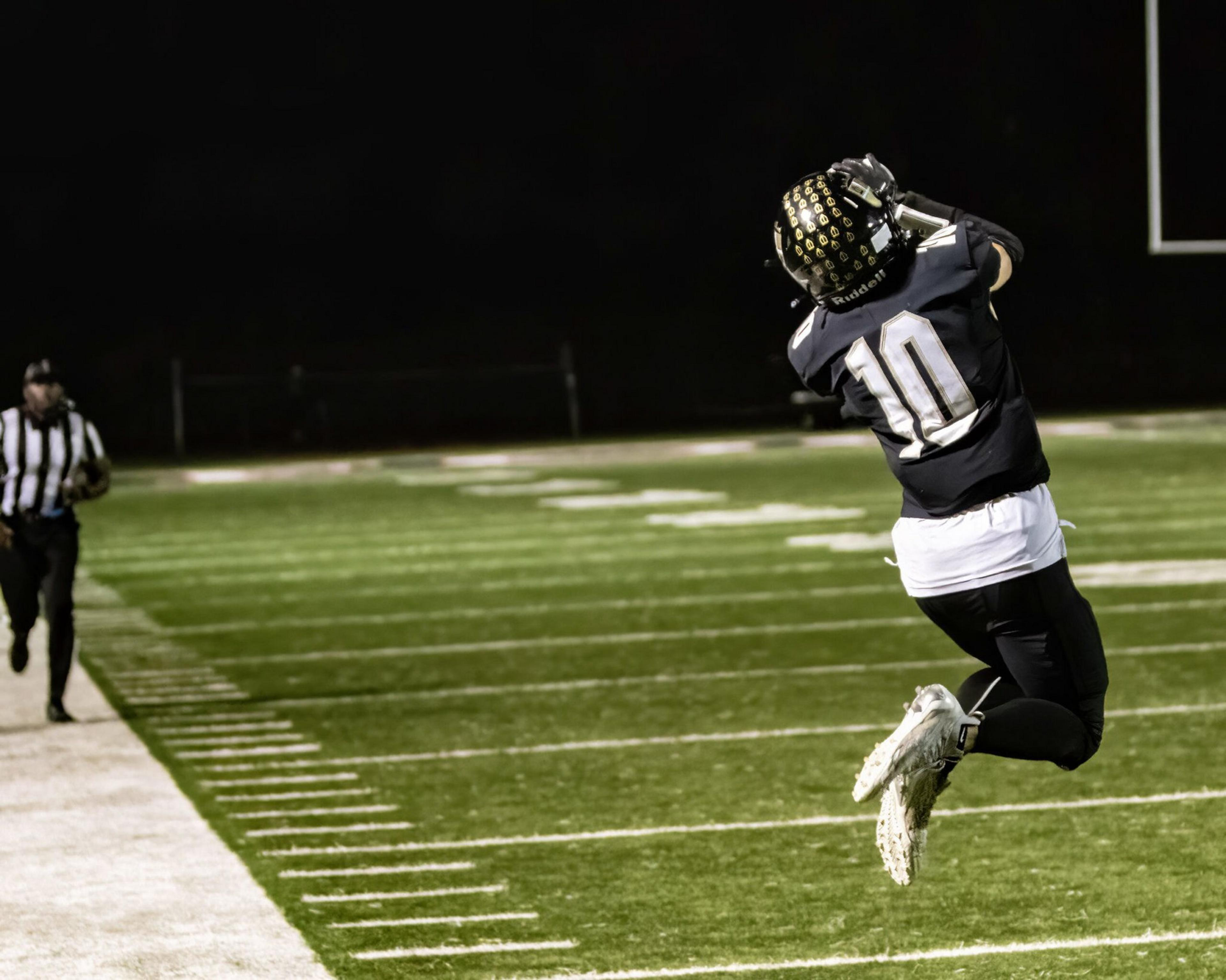 A football player wearing a black jersey with the number 10 leaps to catch a ball near the sideline under stadium lights at night.
