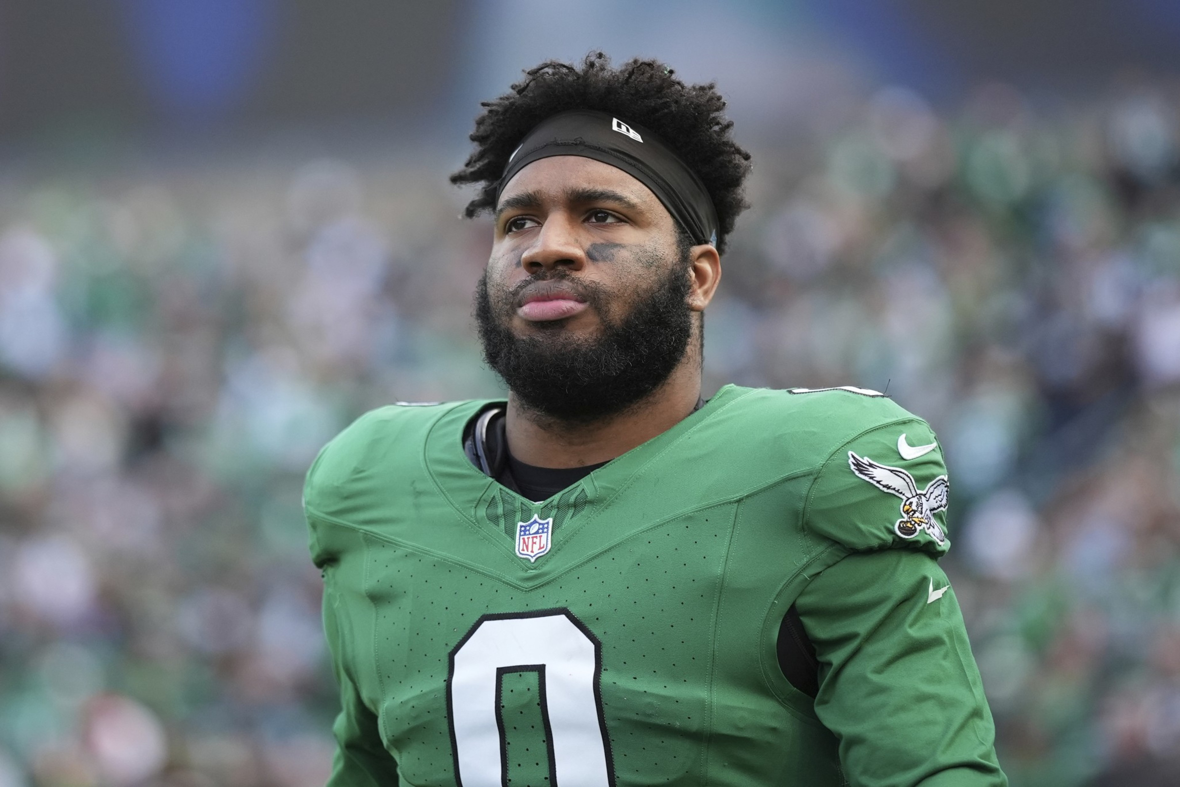 A football player wearing a green jersey with the number 0 and a headband stands on a field. The background has a crowd, creating a blurred effect.