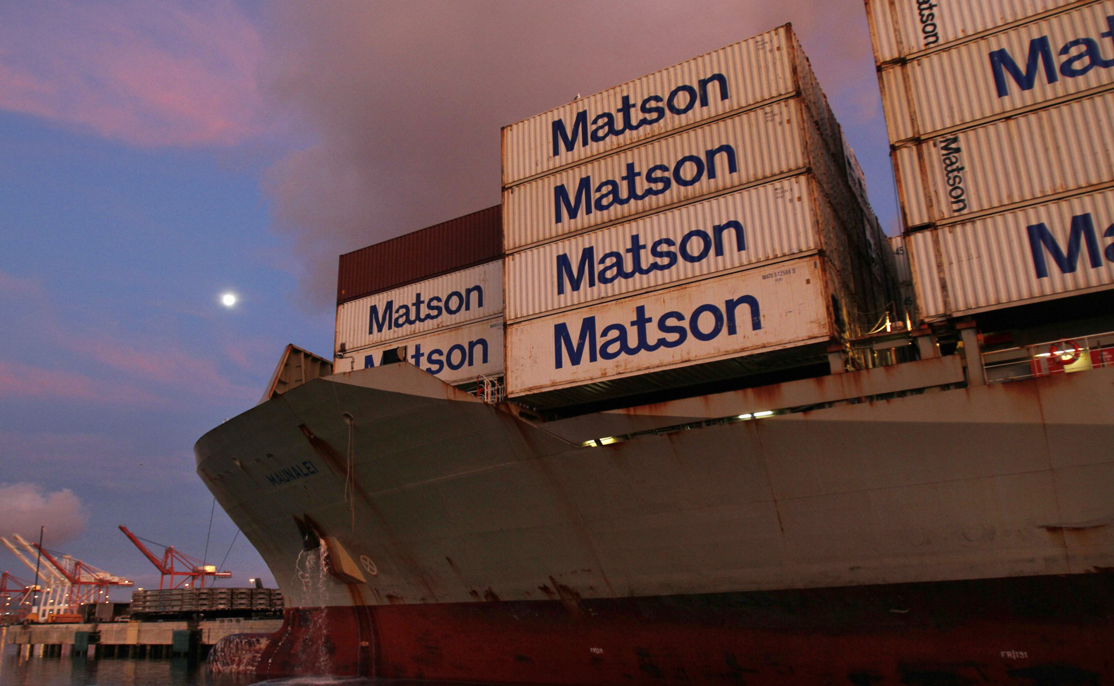 A cargo ship loaded with stacked Matson shipping containers sits docked at a port during sunset under a partly cloudy sky with a visible moon.