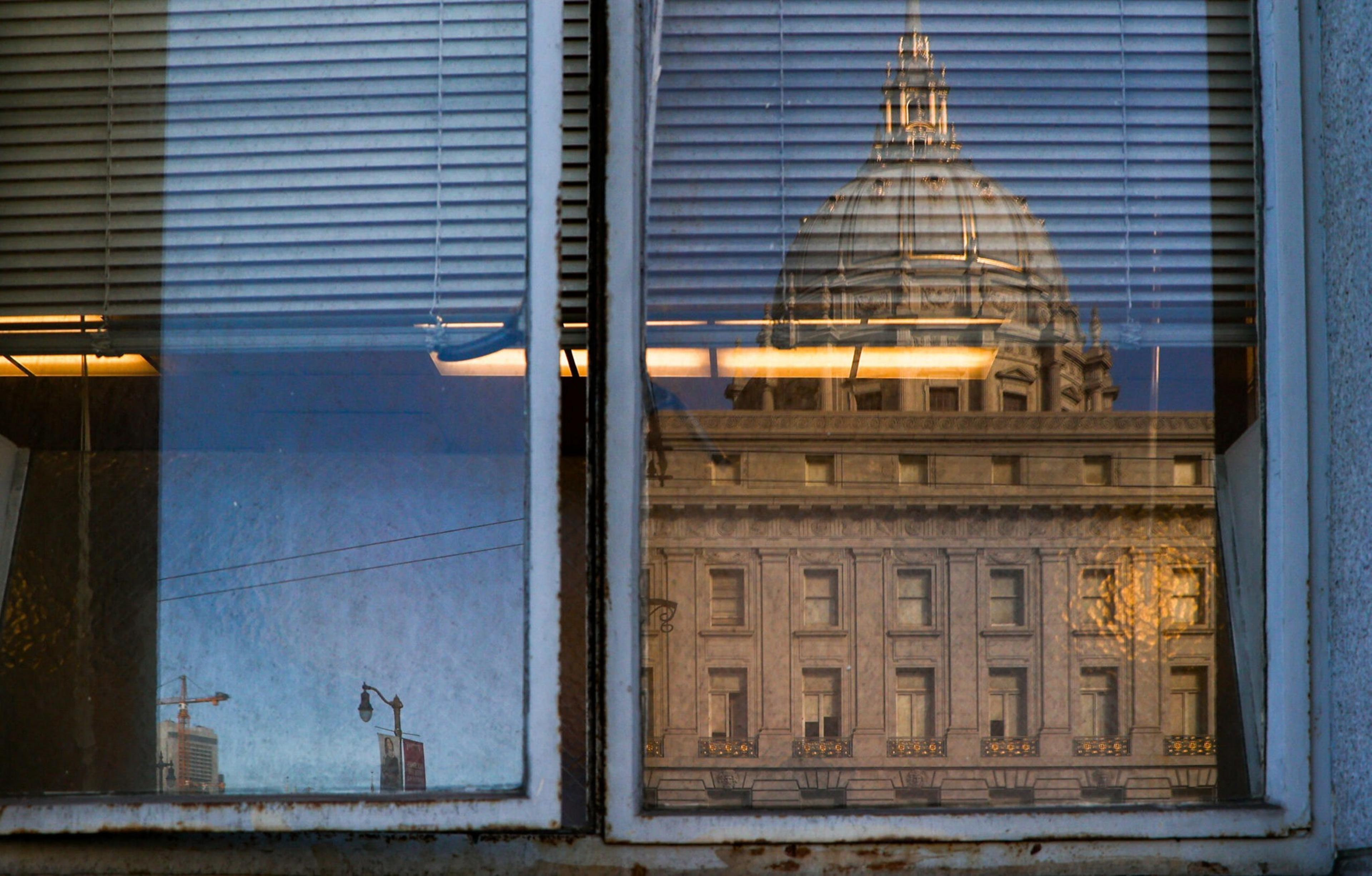 A large historic building with a dome is reflected in an old window with partly closed blinds and a rusty frame.