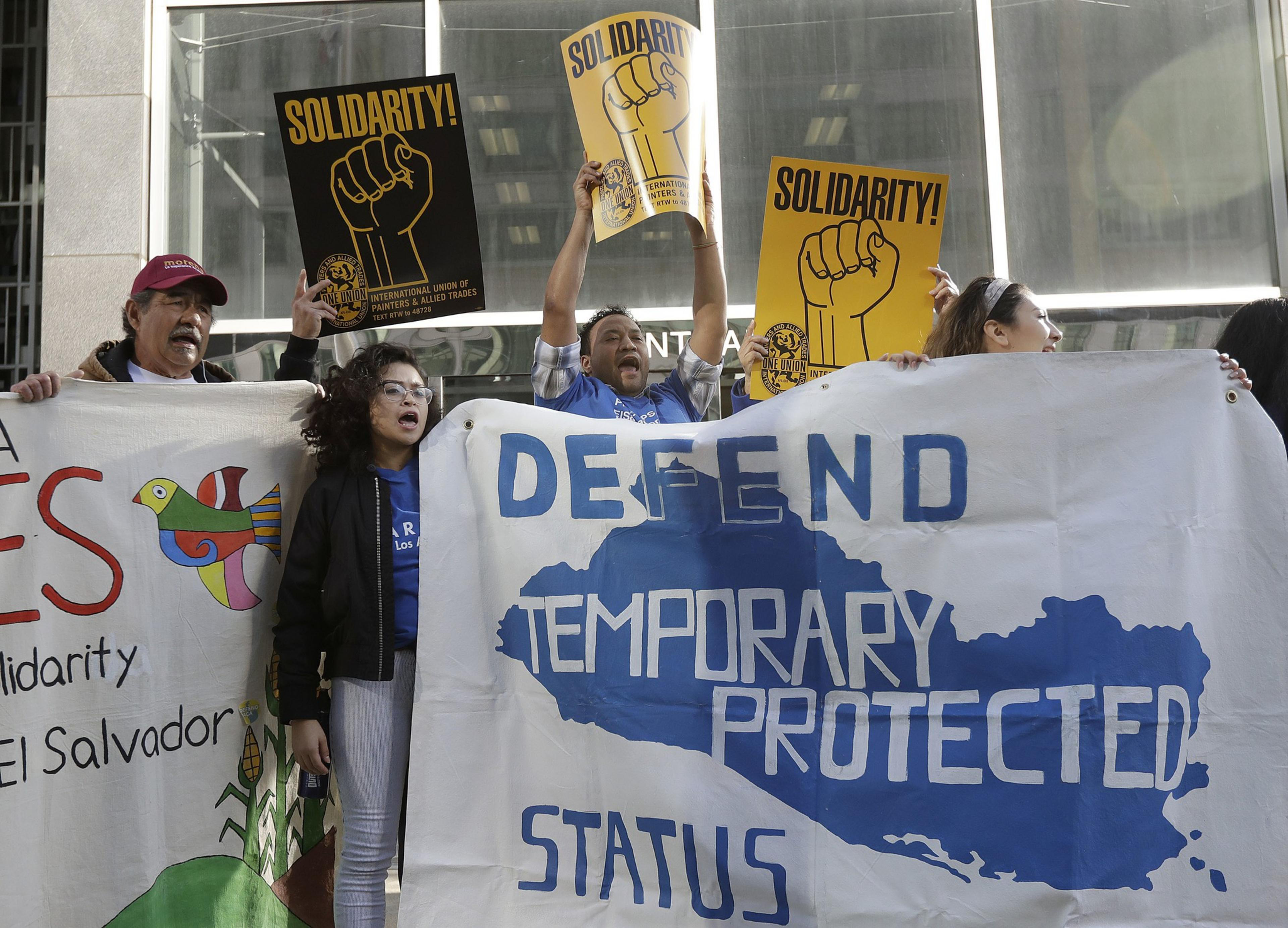 Protesters hold signs reading &quot;Solidarity!&quot; and a banner saying &quot;Defend Temporary Protected Status&quot; in bold text, amidst a backdrop of a building.