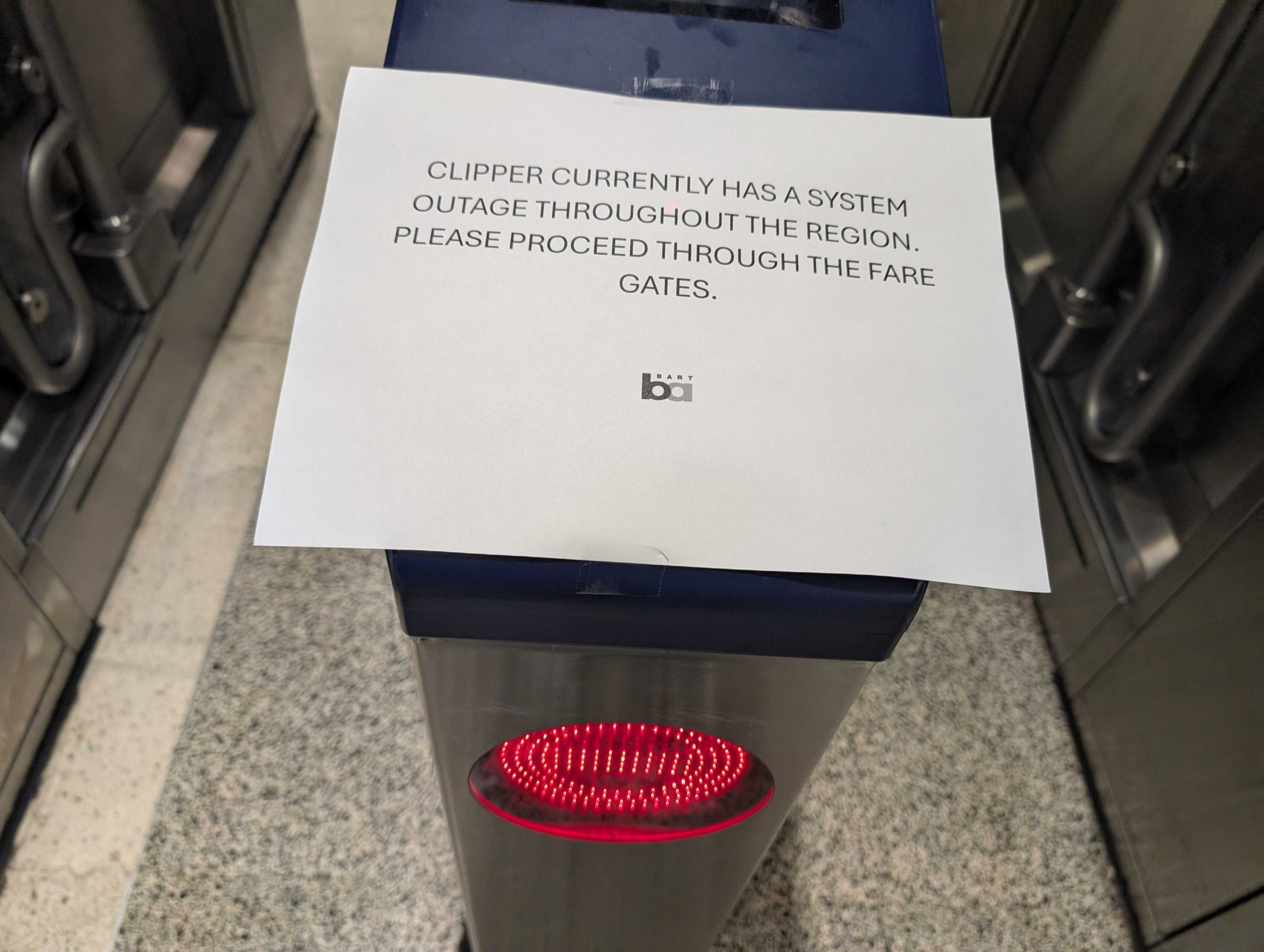 A sign on a transit fare gate reads, &quot;Clipper currently has a system outage throughout the region. Please proceed through the fare gates.&quot;