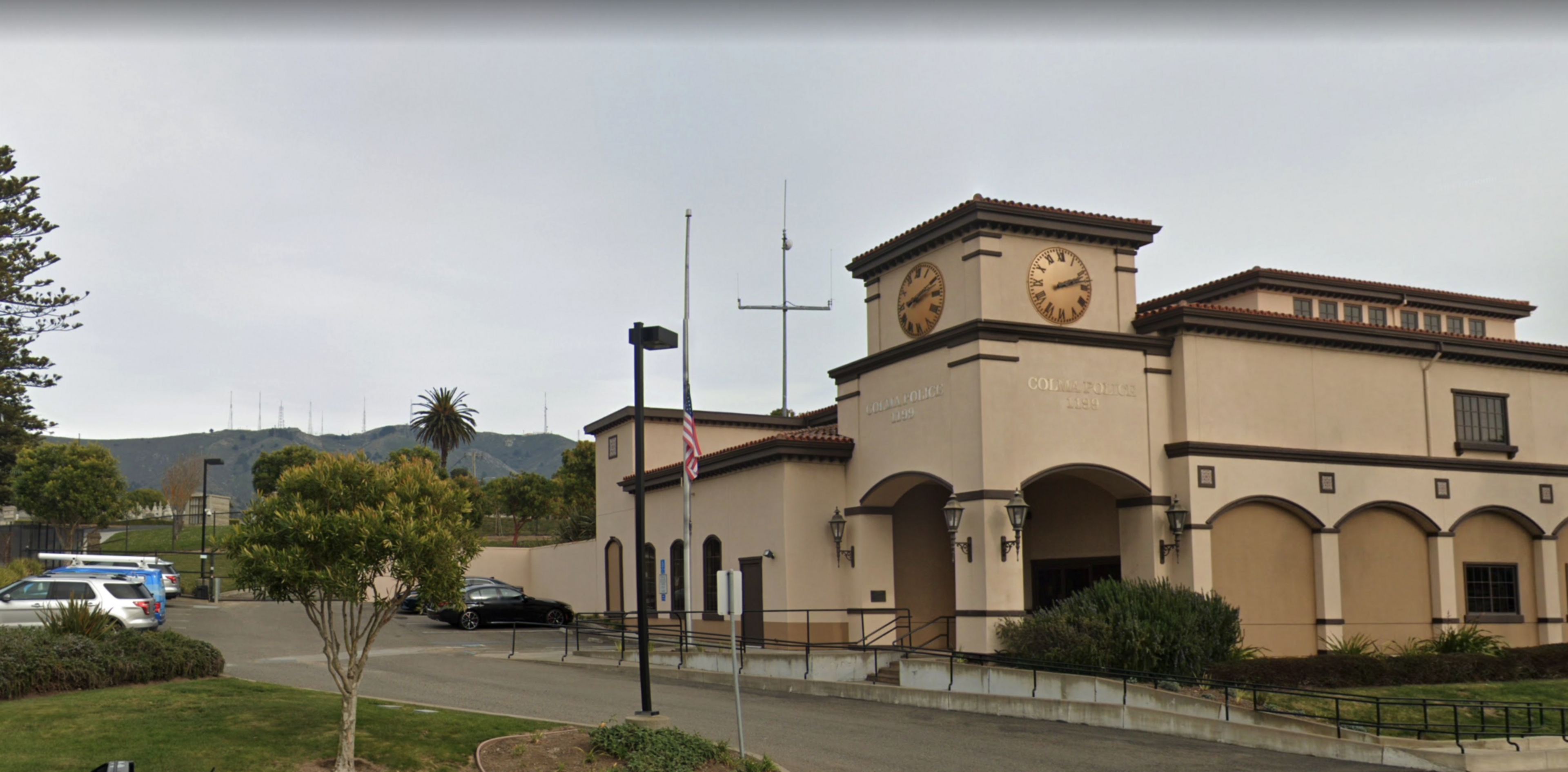 A beige public building with a clock tower shows two clocks reading 11:05 and 1:25, with an American flag flying at half-mast in front.