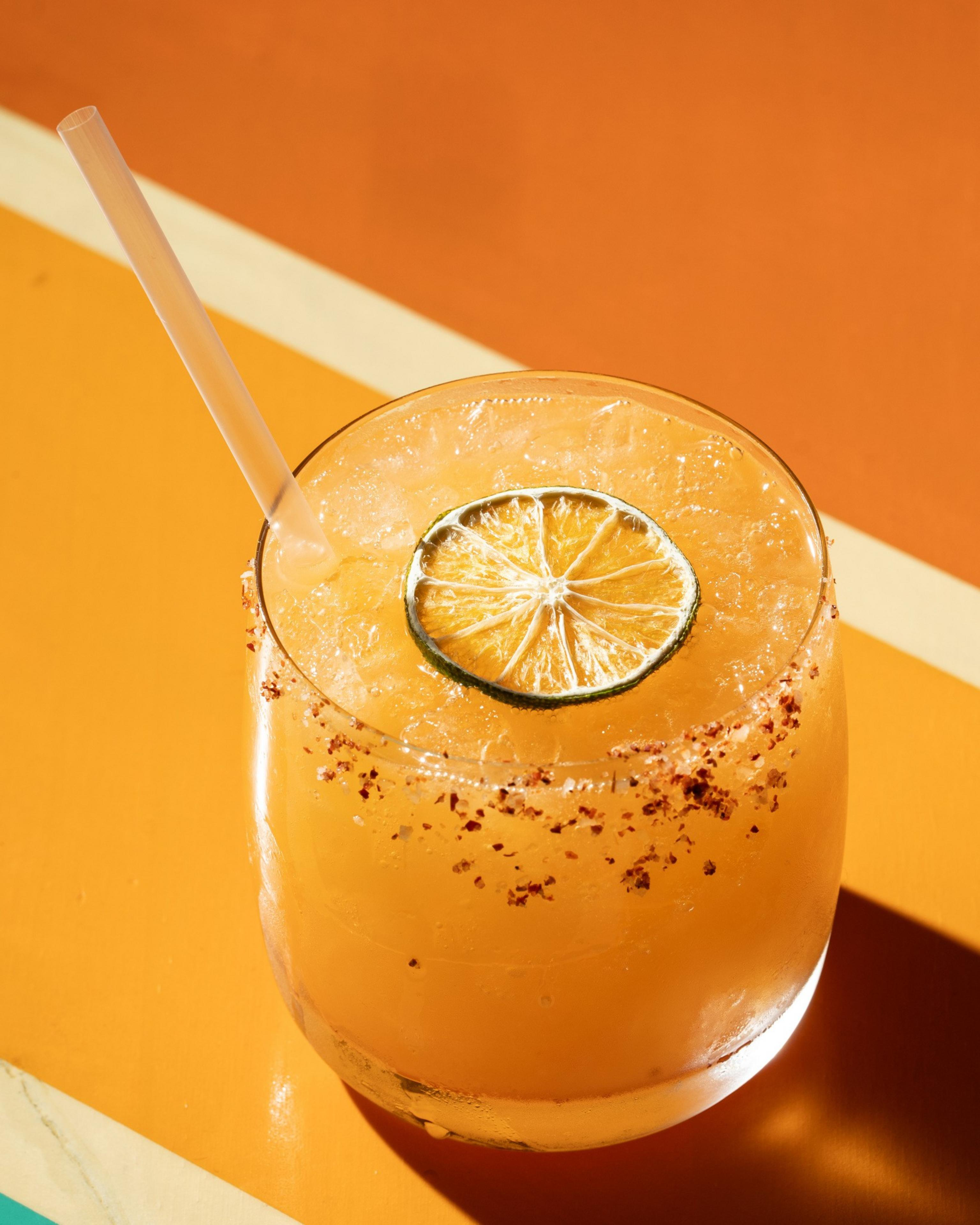 A refreshing orange drink garnished with a lime slice and a chili powder-rimmed edge sits on an orange and yellow surface, featuring a straw.