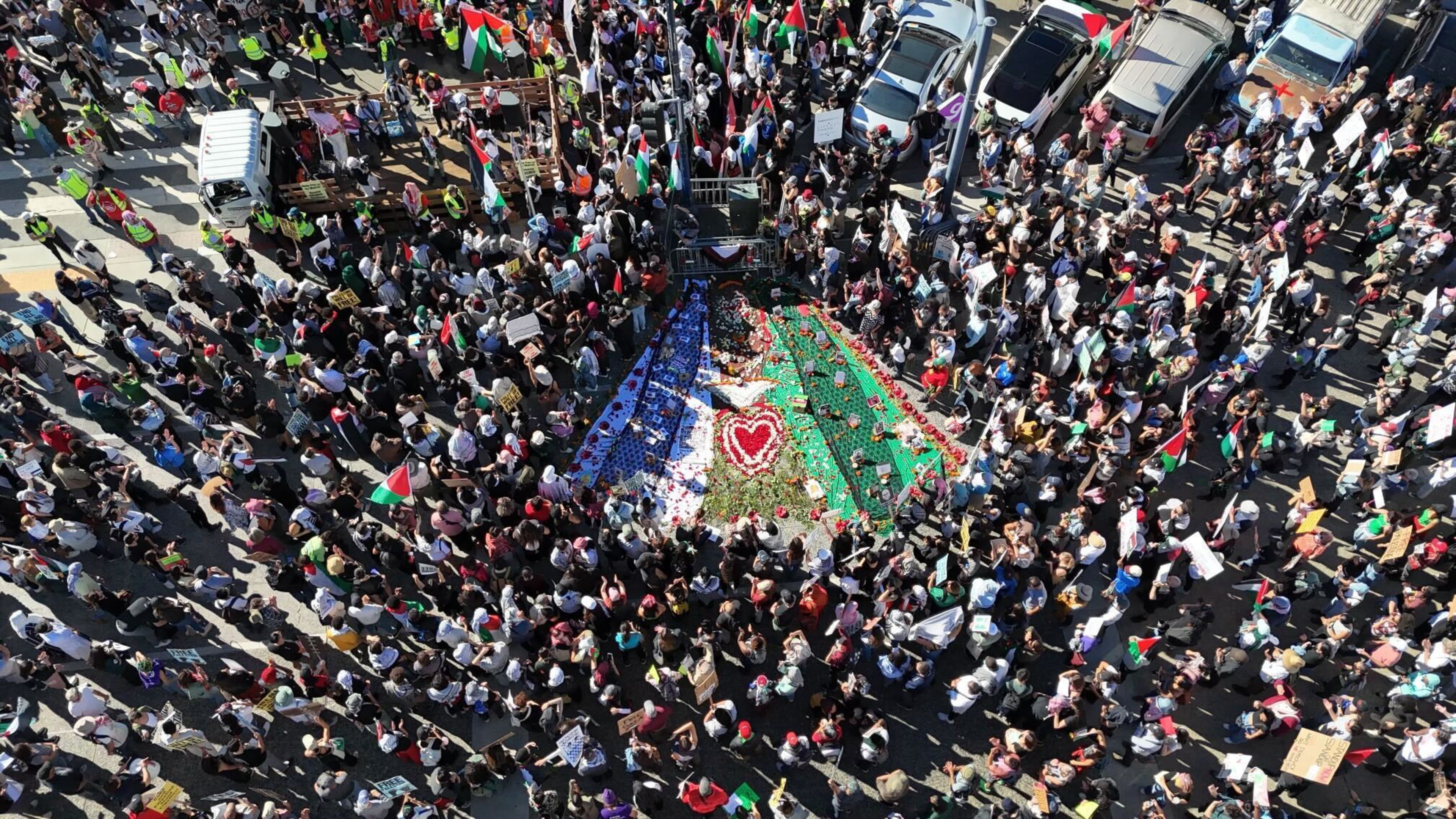 A large crowd surrounds a ground mural resembling the Palestinian flag with flowers and a red heart in the center, many holding Palestinian flags.