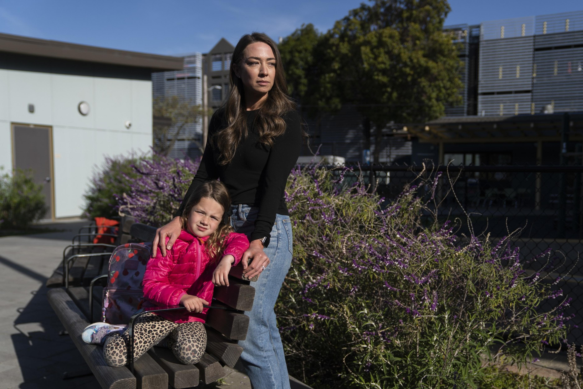 A woman in a black top and jeans stands next to a sitting child in a pink jacket and leopard-print leggings. They're outdoors near purple flowers.