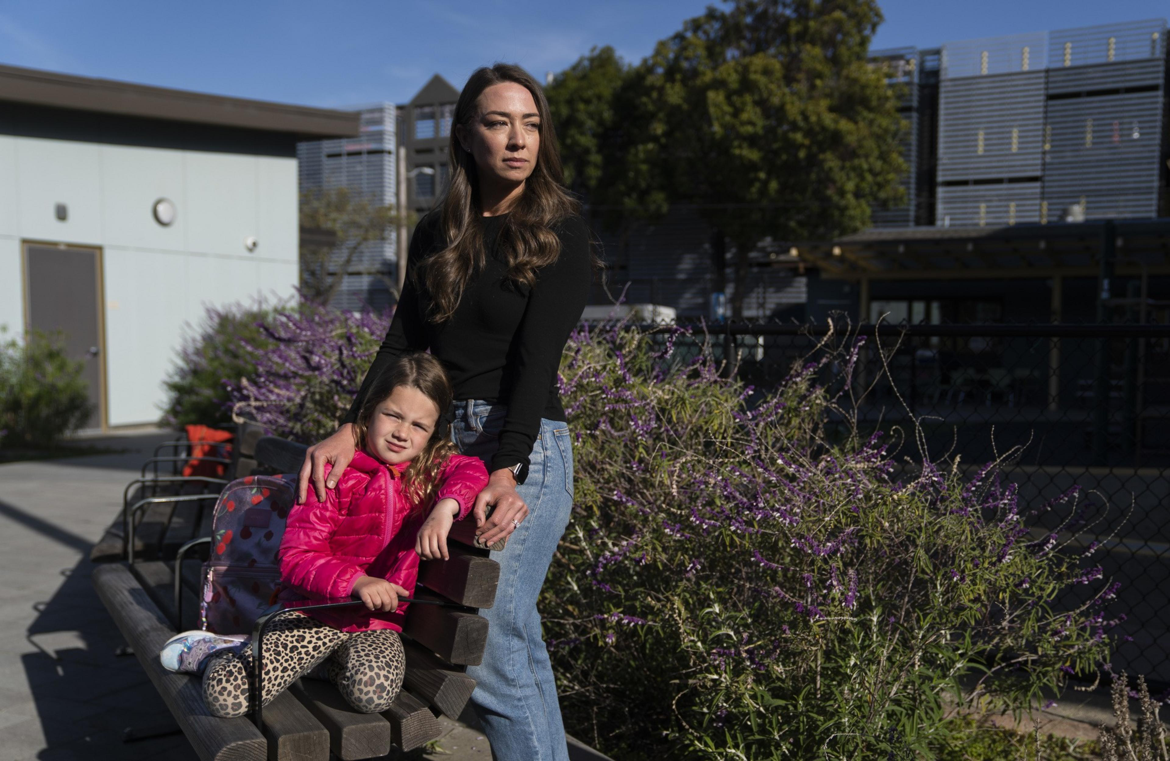 A woman in a black top and jeans stands next to a sitting child in a pink jacket and leopard-print leggings. They're outdoors near purple flowers.