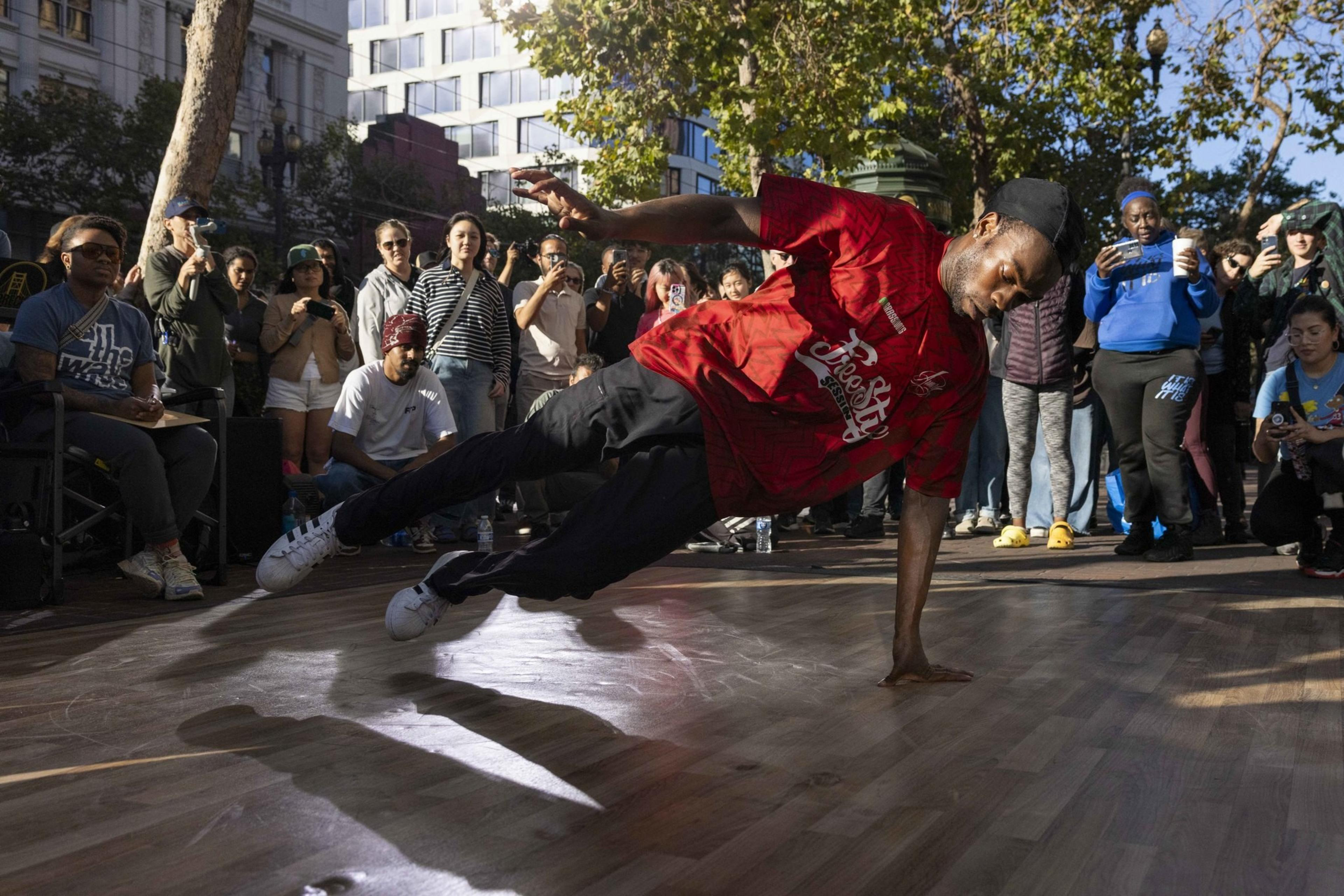 A street dancer in a red shirt performs a dynamic move on a wooden floor, while a crowd of onlookers, some filming, gathers around in an outdoor urban setting.