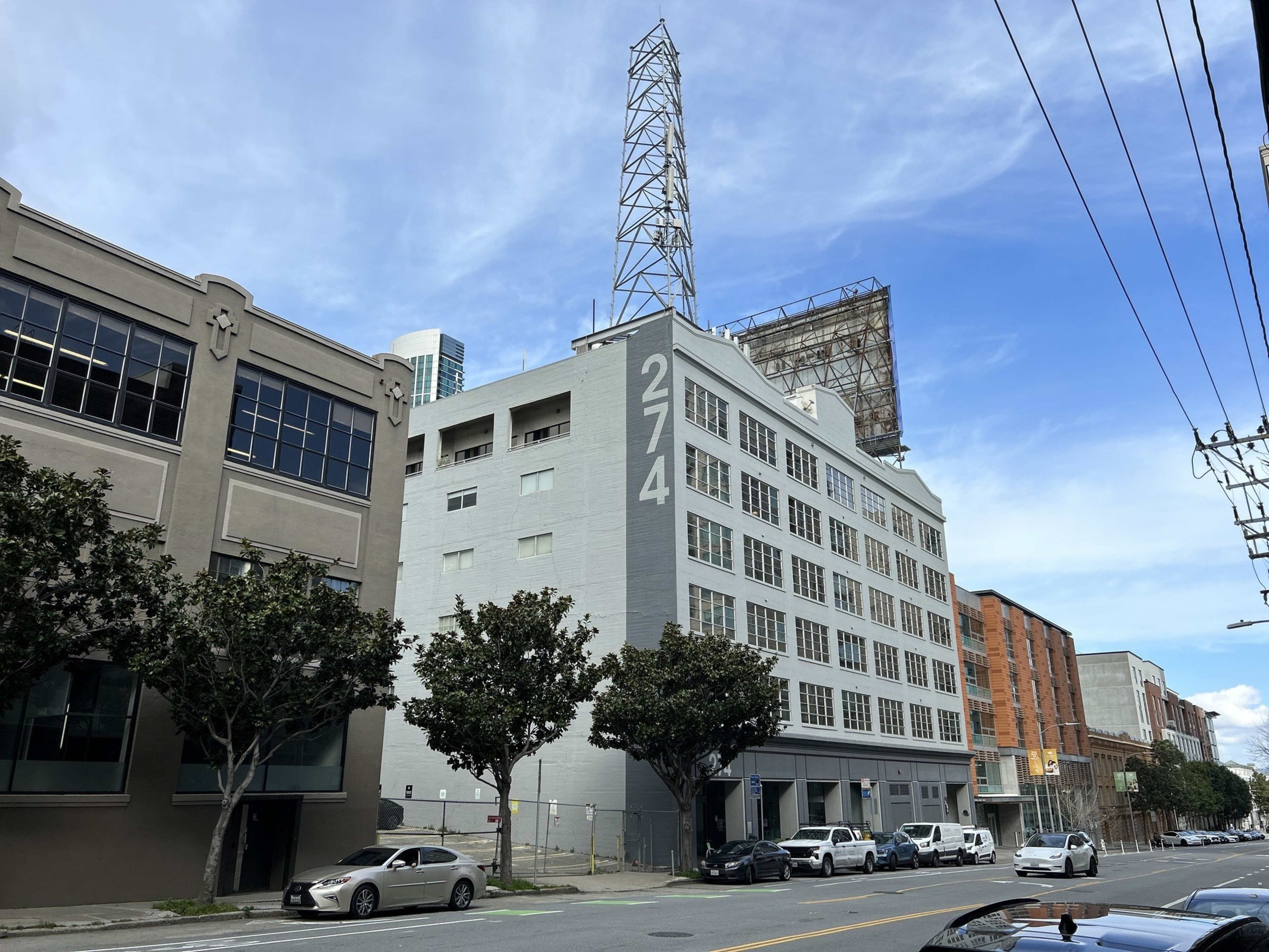 Urban street with multi-story buildings, a large &quot;274&quot; painted on one, a tall metal tower, and parked cars under a blue sky.
