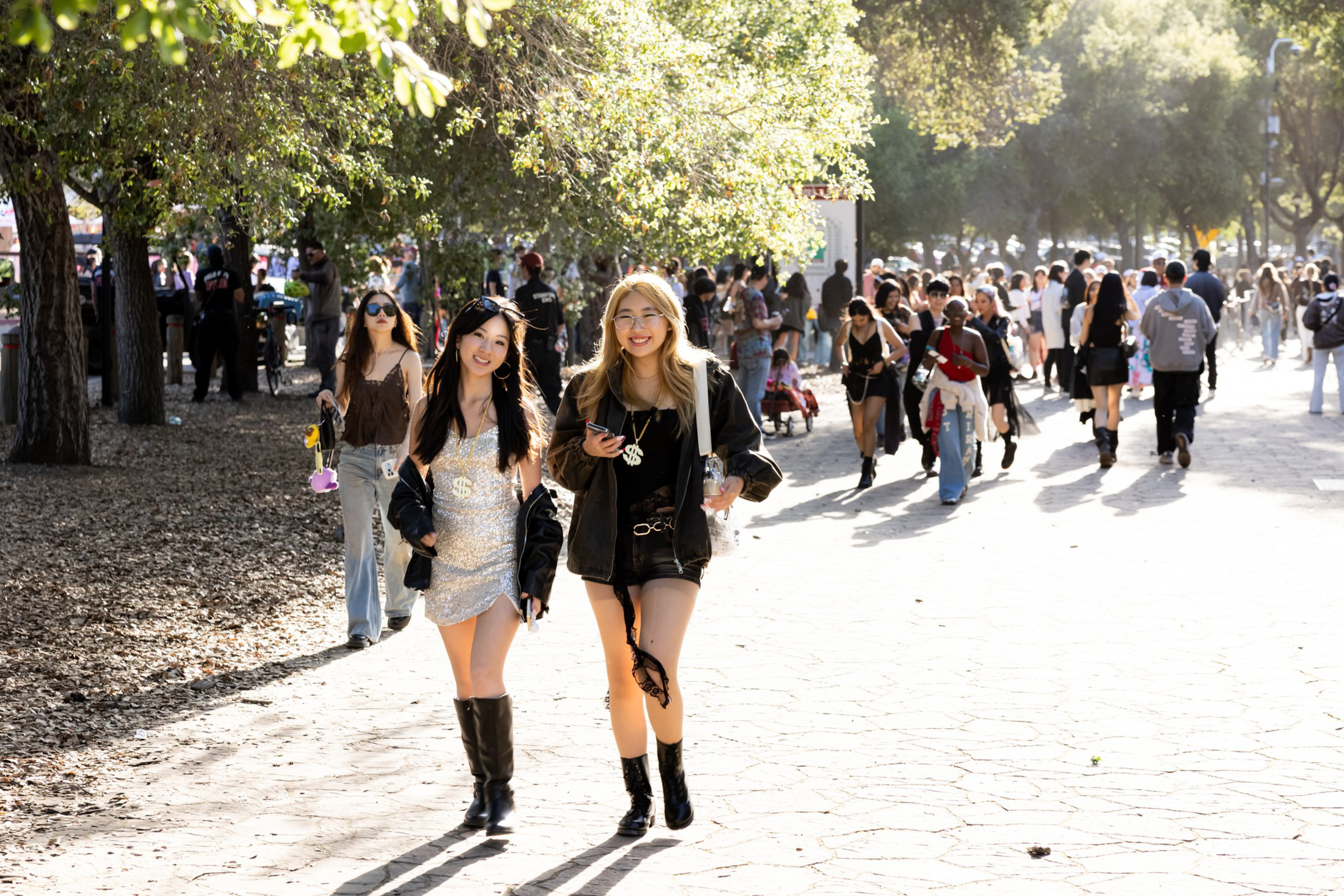 Two young women walk smiling on a sunlit path, wearing stylish outfits with boots, while a crowd of people strolls in the background amidst trees.