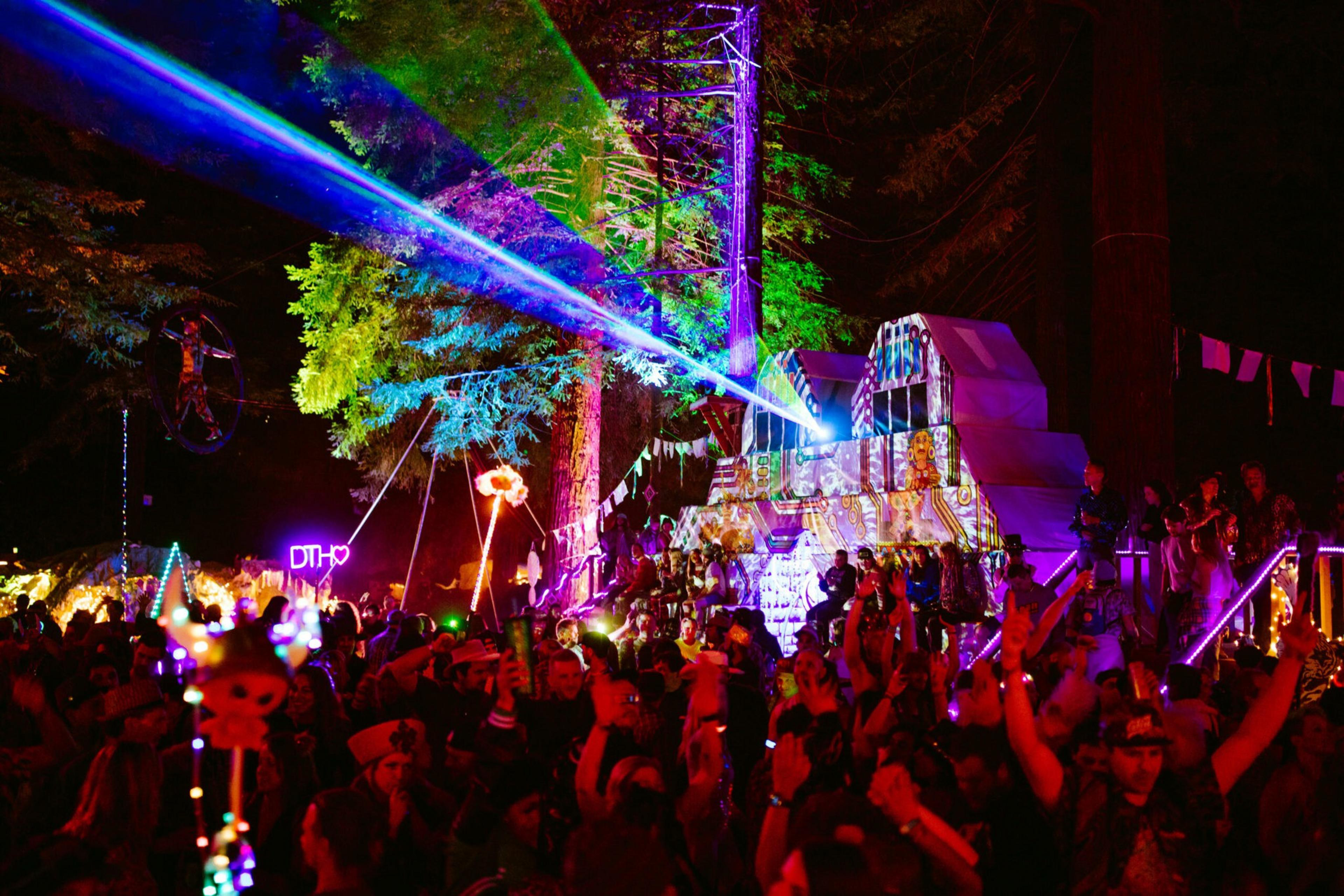 A vibrant crowd dances at night under colorful laser lights beaming from a decorated stage surrounded by large trees and illuminated decorations.