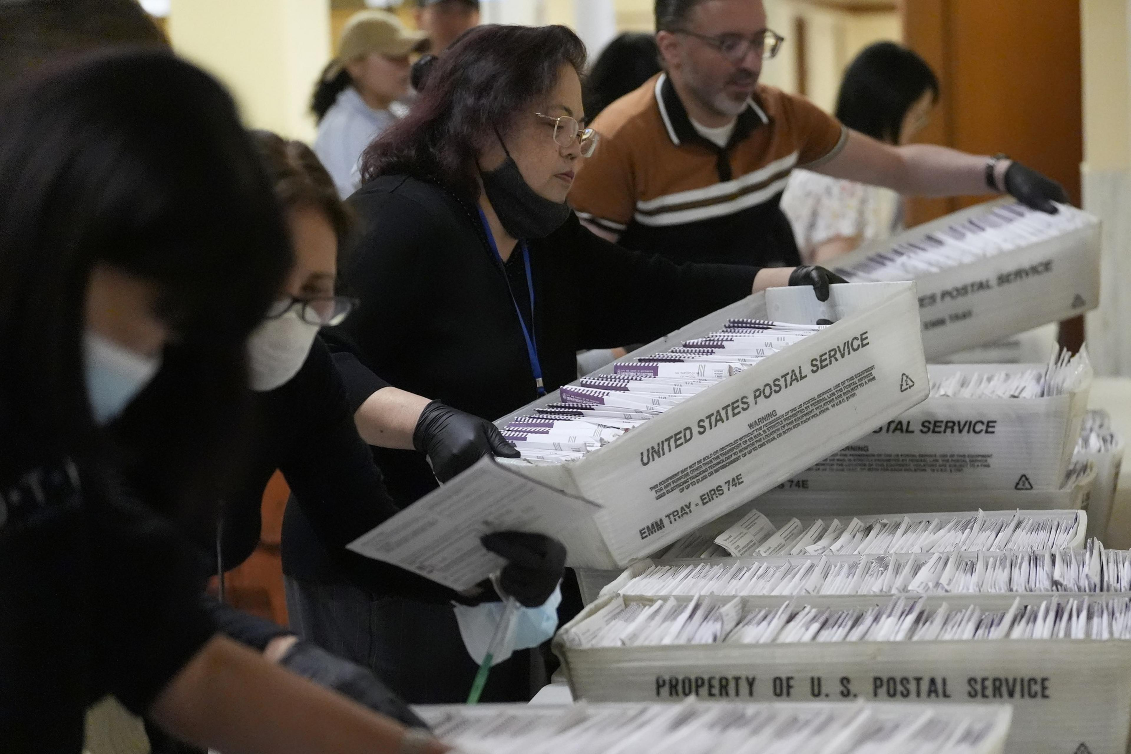 People are sorting and handling stacks of envelopes in USPS trays, likely mail ballots. They wear masks and gloves, working closely together.