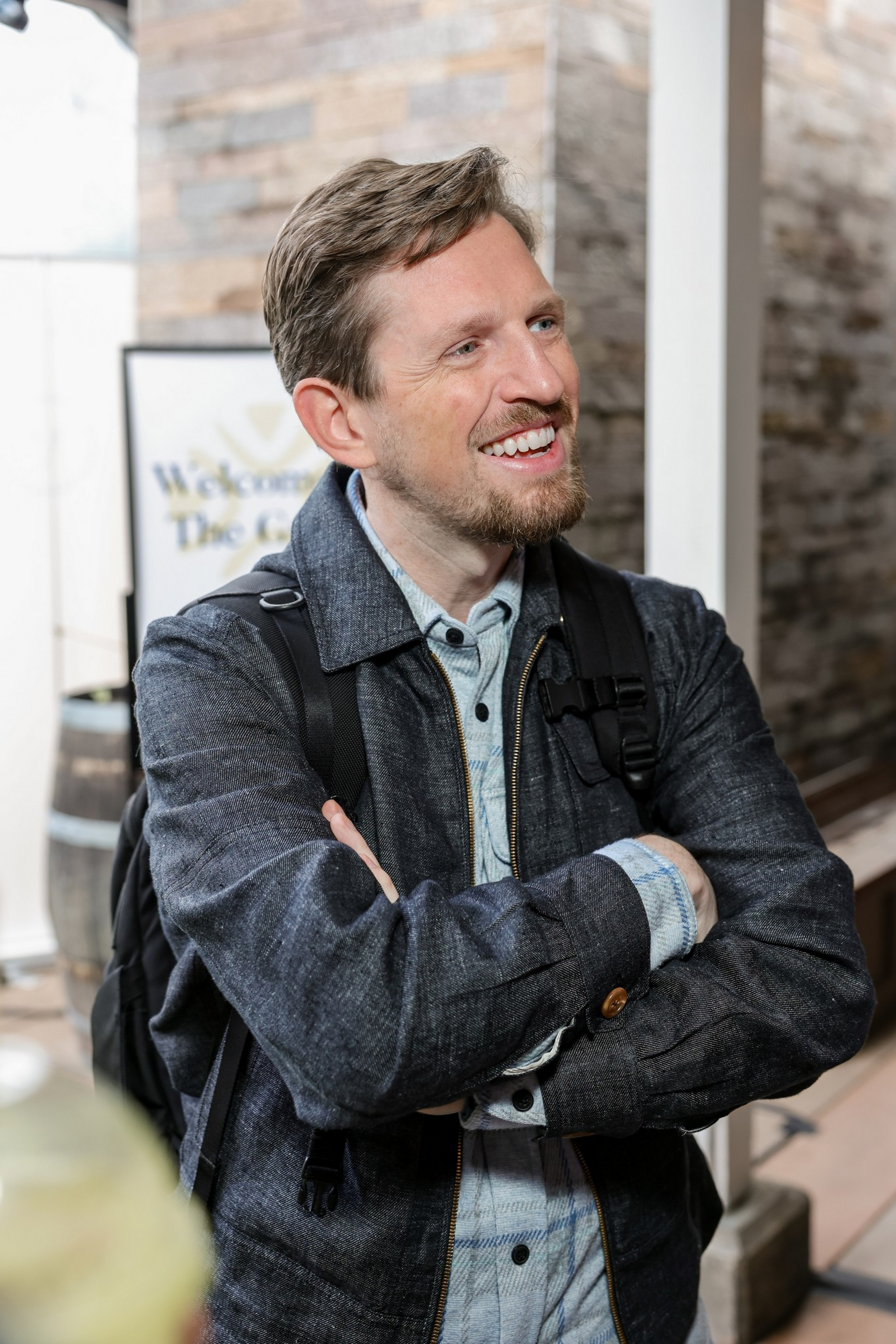 A smiling man with light brown hair and a beard wears a dark jacket and backpack, standing with arms crossed indoors next to a wooden barrel and a sign.