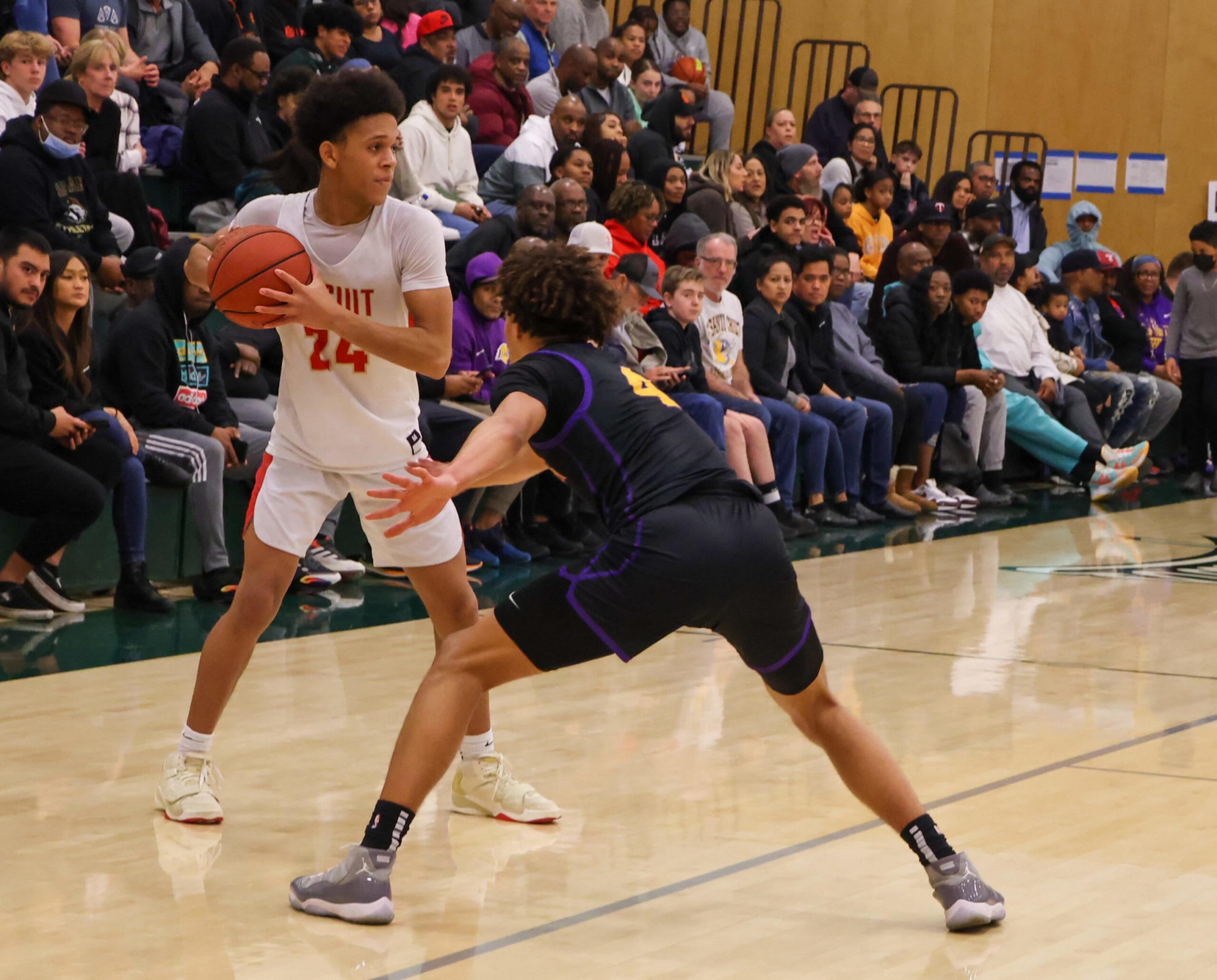 A basketball player in a white jersey holds the ball, guarded closely by a player in a black jersey, with a crowd watching from the bleachers.
