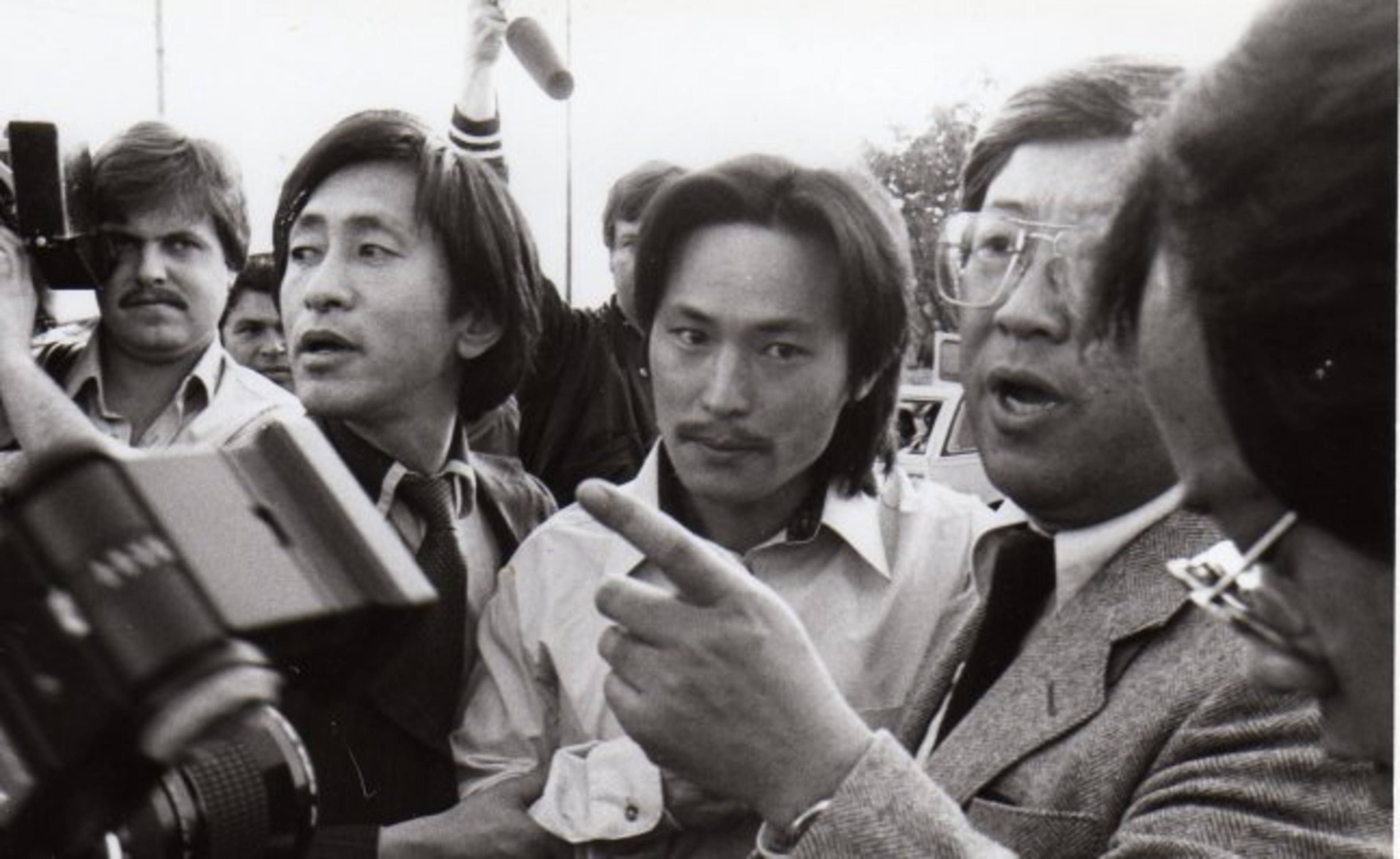 A group of men, some with cameras, crowd closely as one man in glasses points and speaks, appearing to address or direct the others.