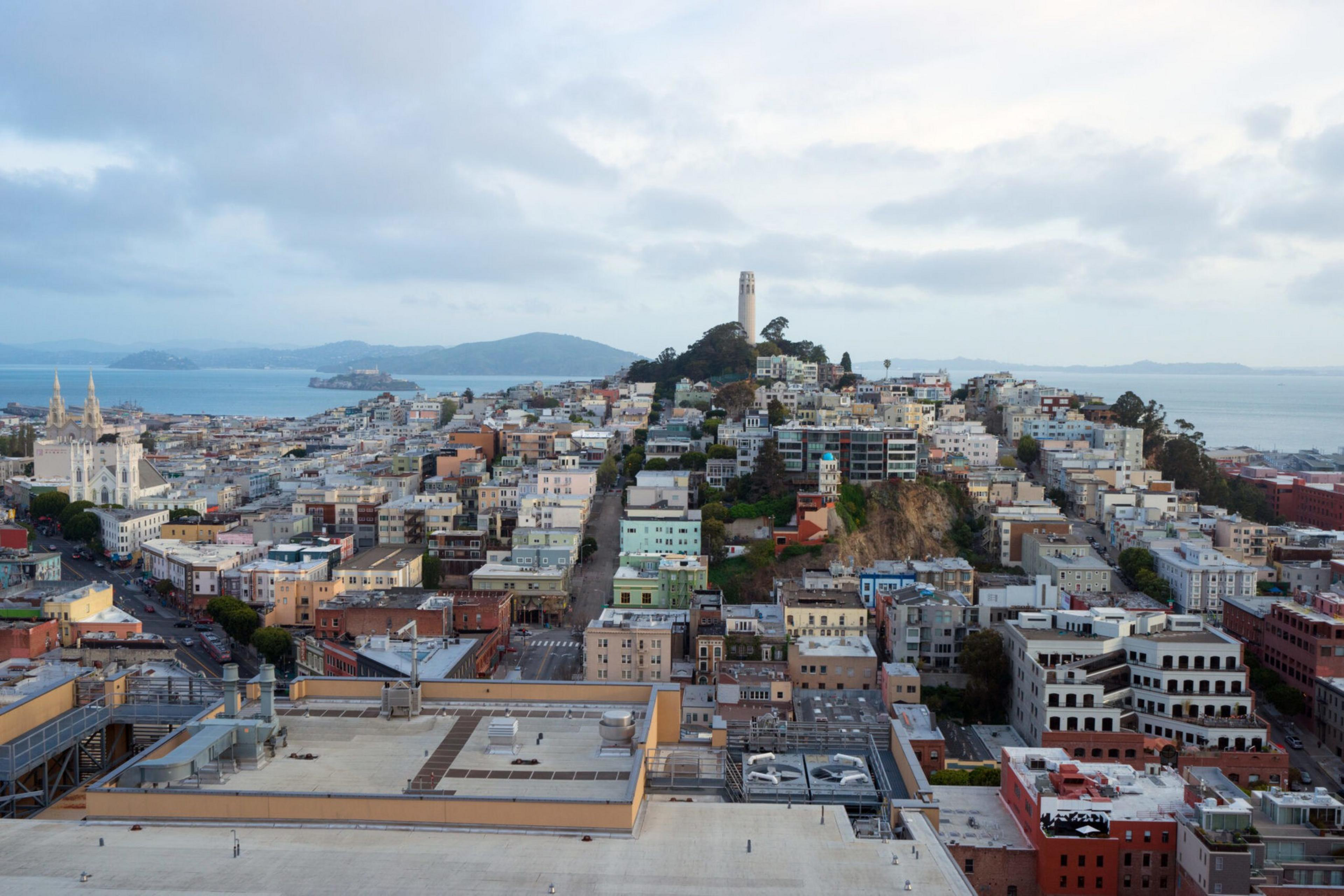 A densely packed cityscape with colorful buildings, a tower on a hill in the center, the bay and distant islands under a cloudy sky in the background.