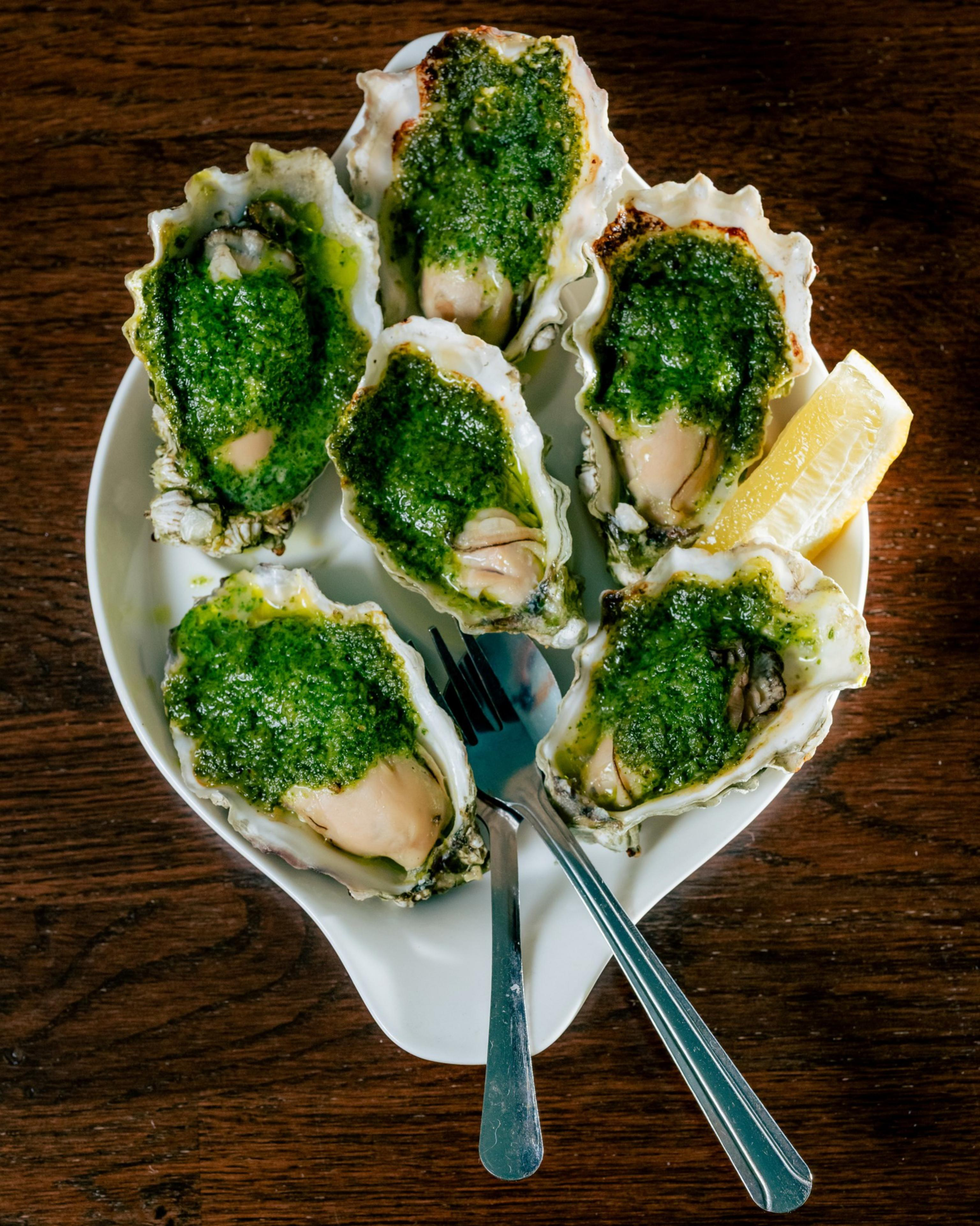 Six oysters topped with green sauce are arranged on a white plate with a lemon wedge and a fork, set on a wooden table.