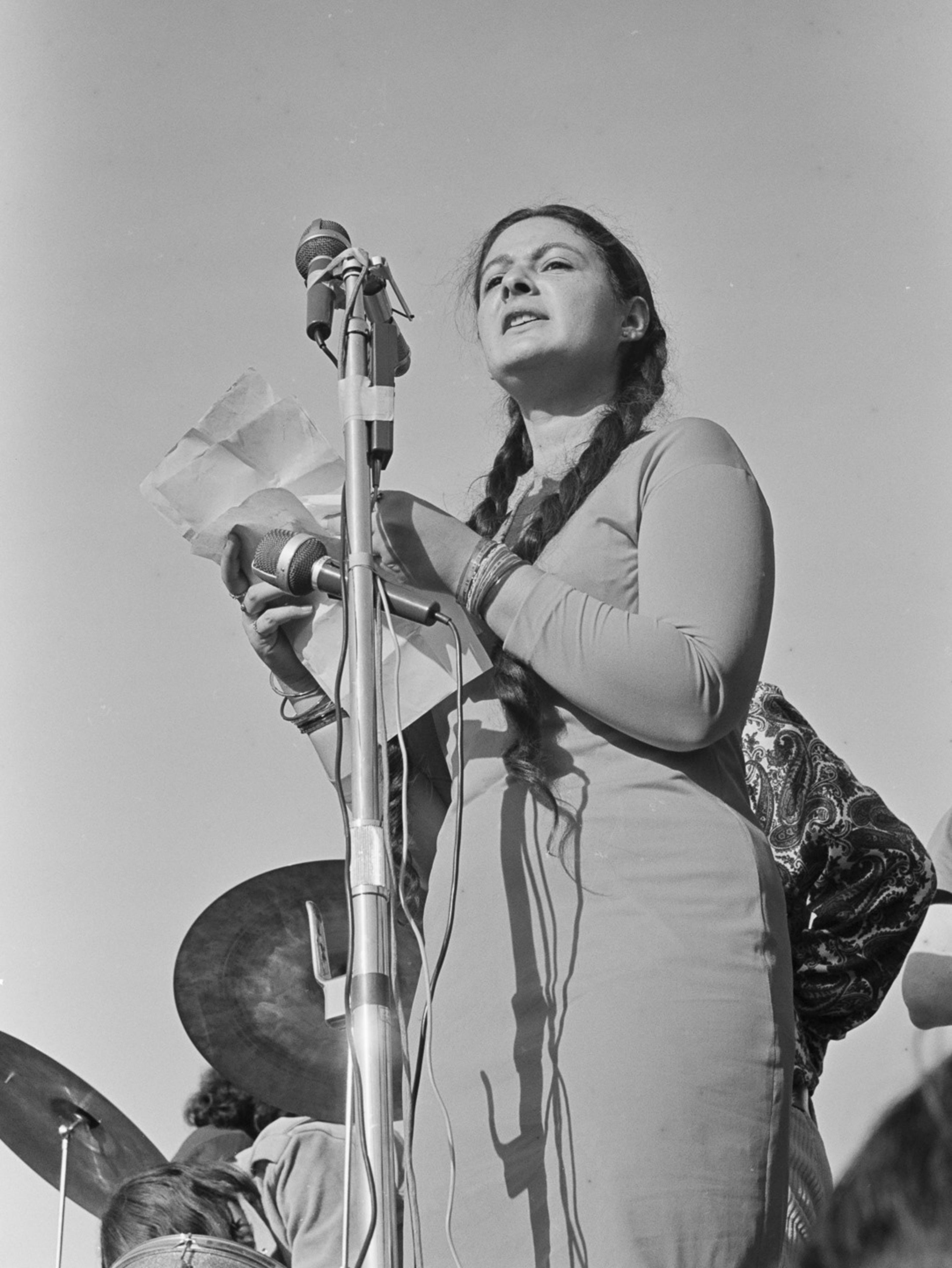 A woman with braided hair is speaking into multiple microphones, holding papers, and standing outdoors under a clear sky, with musical instruments visible behind her.