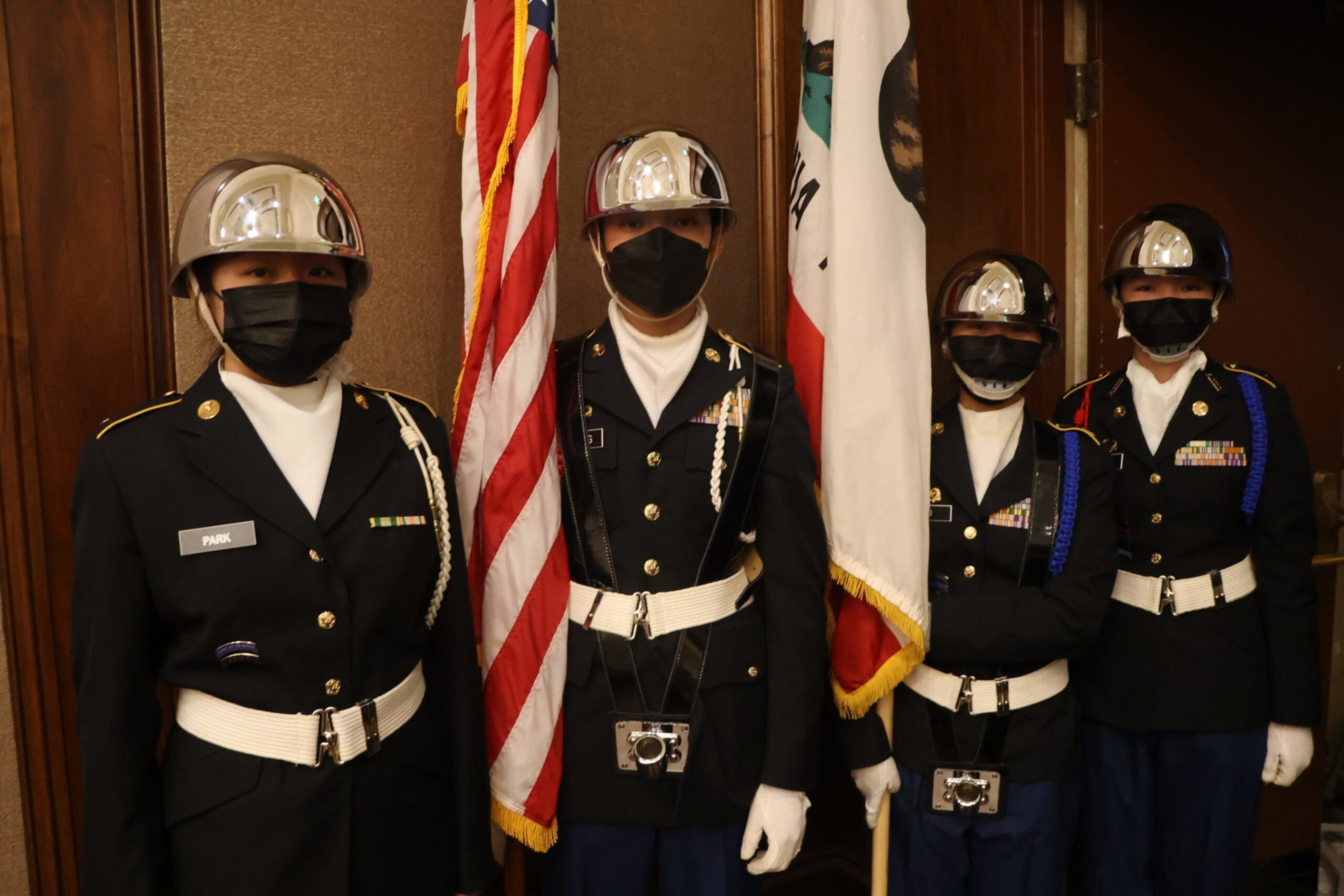 Four uniformed individuals wearing polished helmets and black face masks stand beside American and Californian flags indoors.