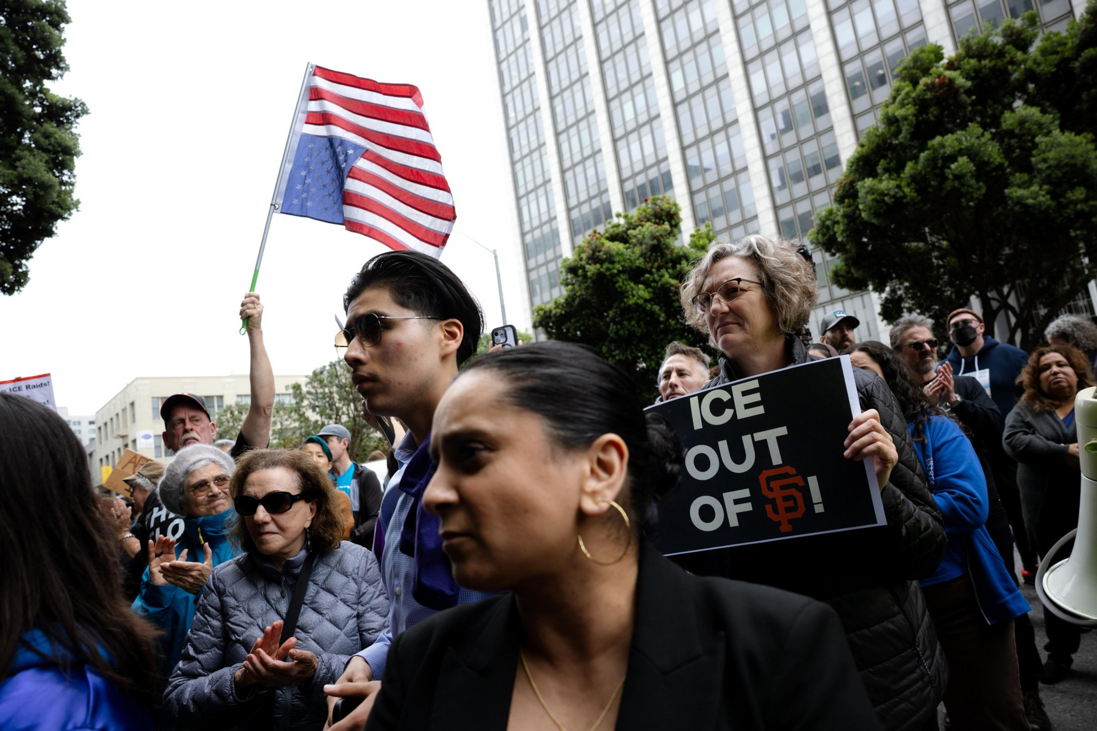 A diverse crowd is protesting outside, holding signs like &quot;ICE OUT OF SF&quot; and an upside-down American flag. They're standing in front of a tall building.