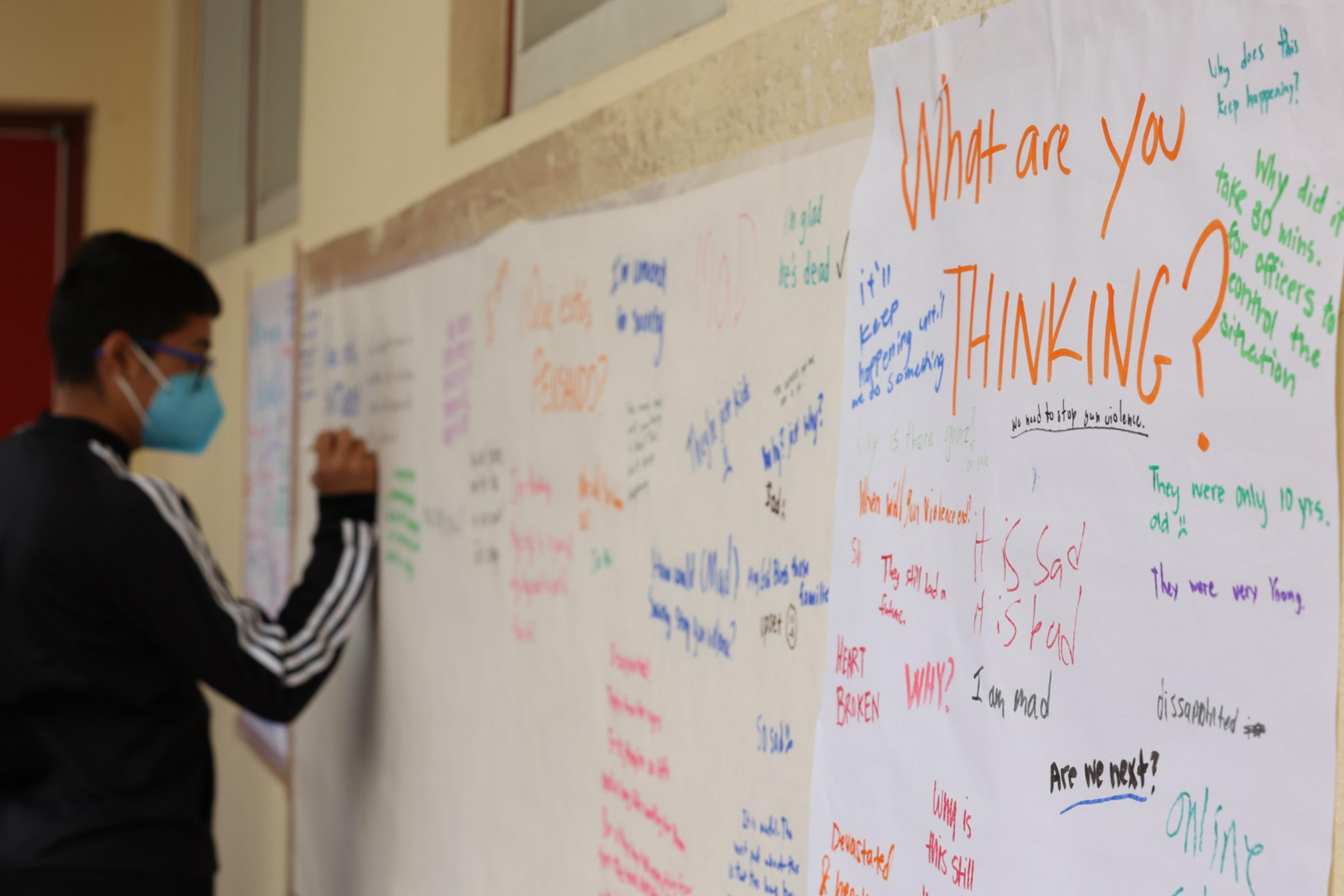 A person wearing a blue mask writes on a wall covered with large white papers filled with colorful handwritten questions and emotional comments.