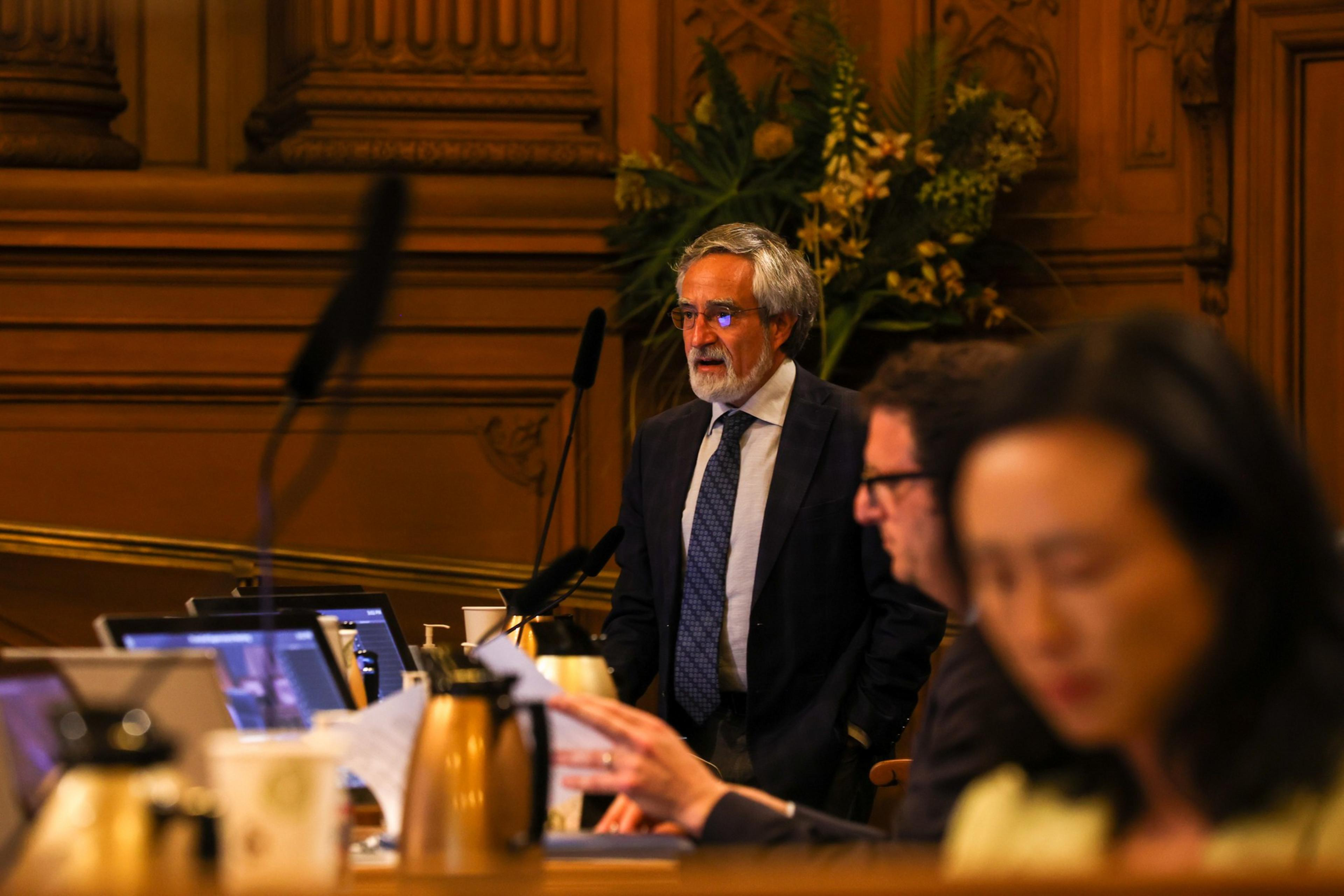 An older man in a suit and tie stands speaking in a wood-paneled room, while two people in the foreground focus on laptops and papers.