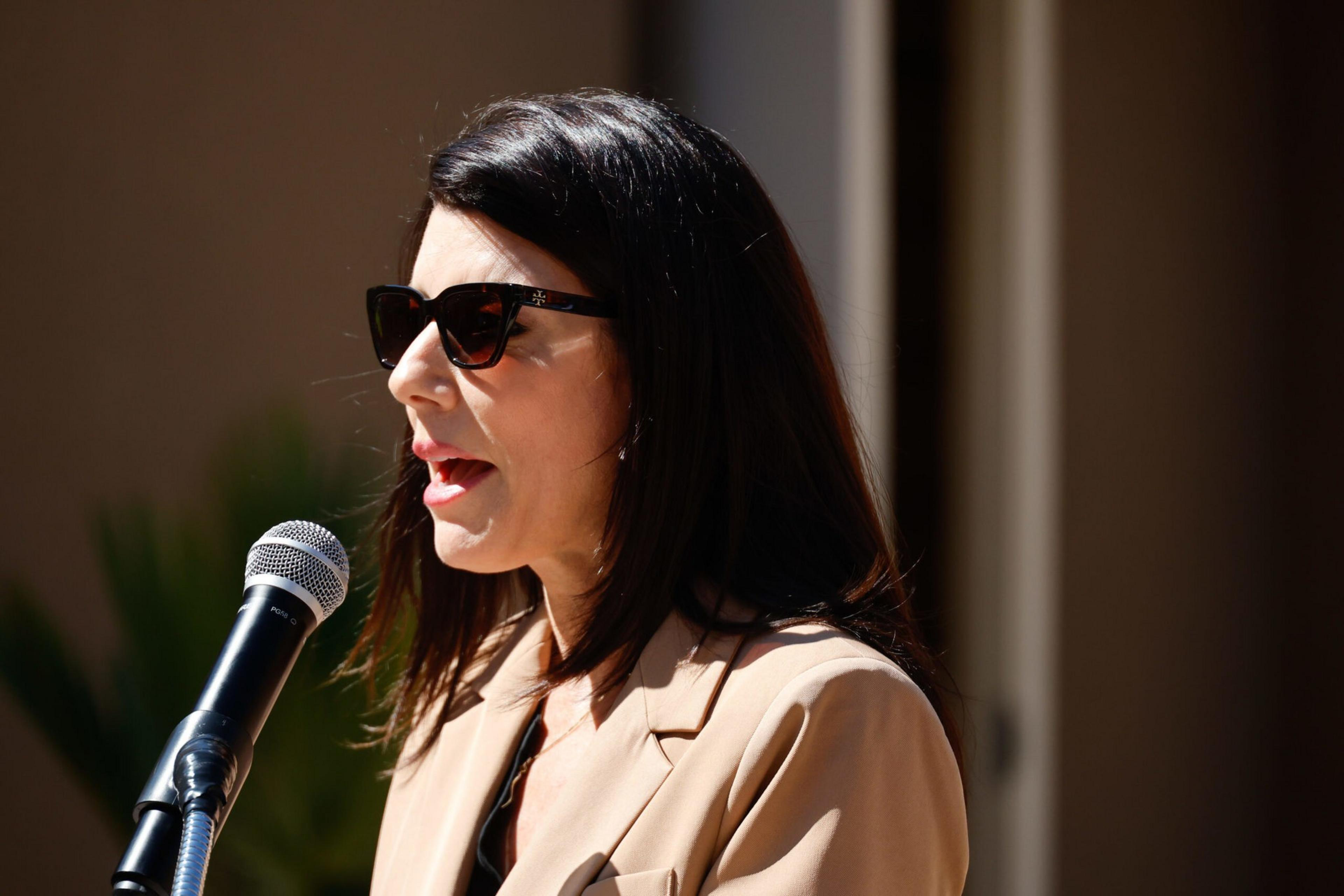A woman with dark hair and sunglasses speaks into a microphone, wearing a beige blazer, against a blurred outdoor background.