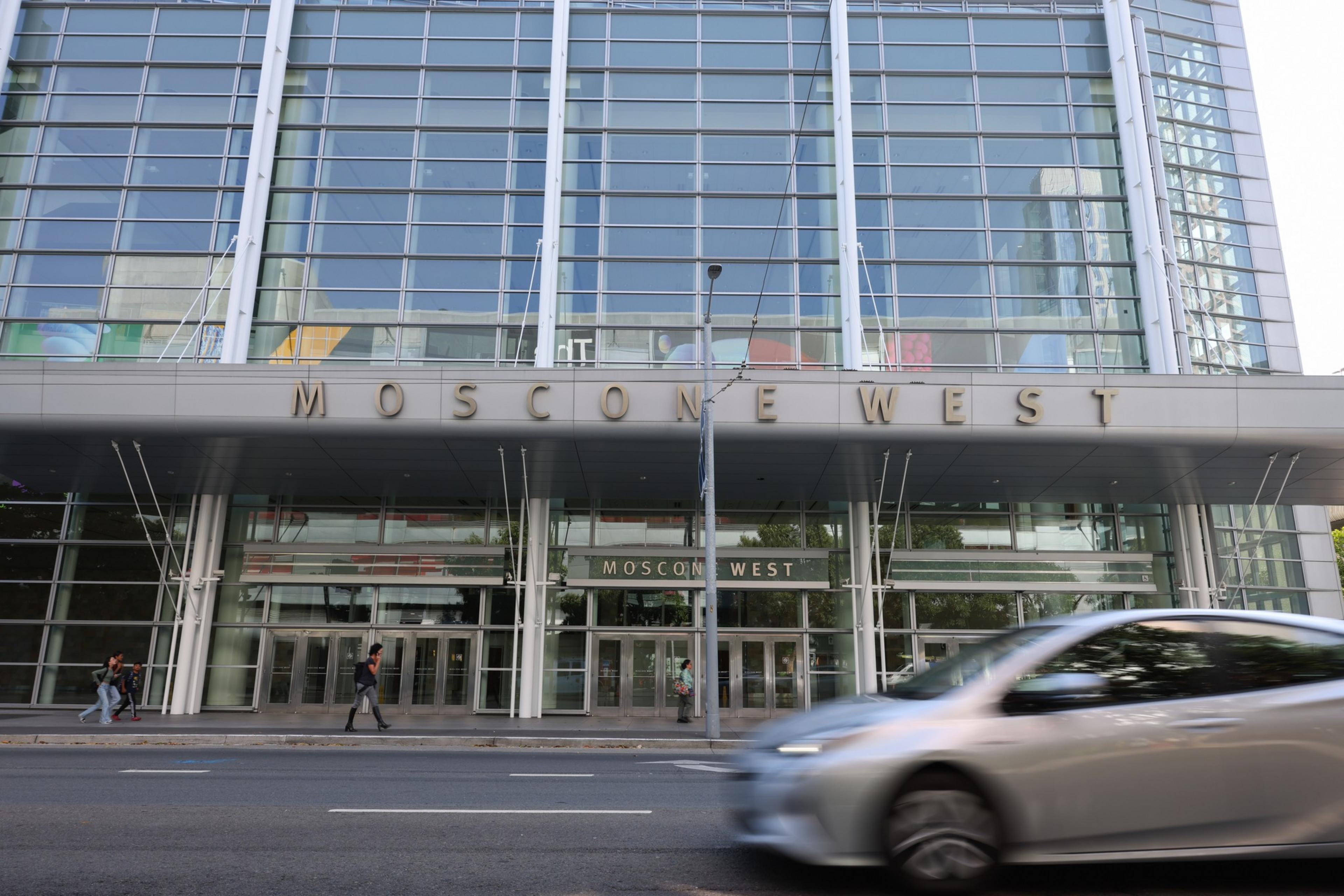 A modern glass building with large windows and the words "Moscone West" above the entrance, with a silver car passing by and a few people walking.