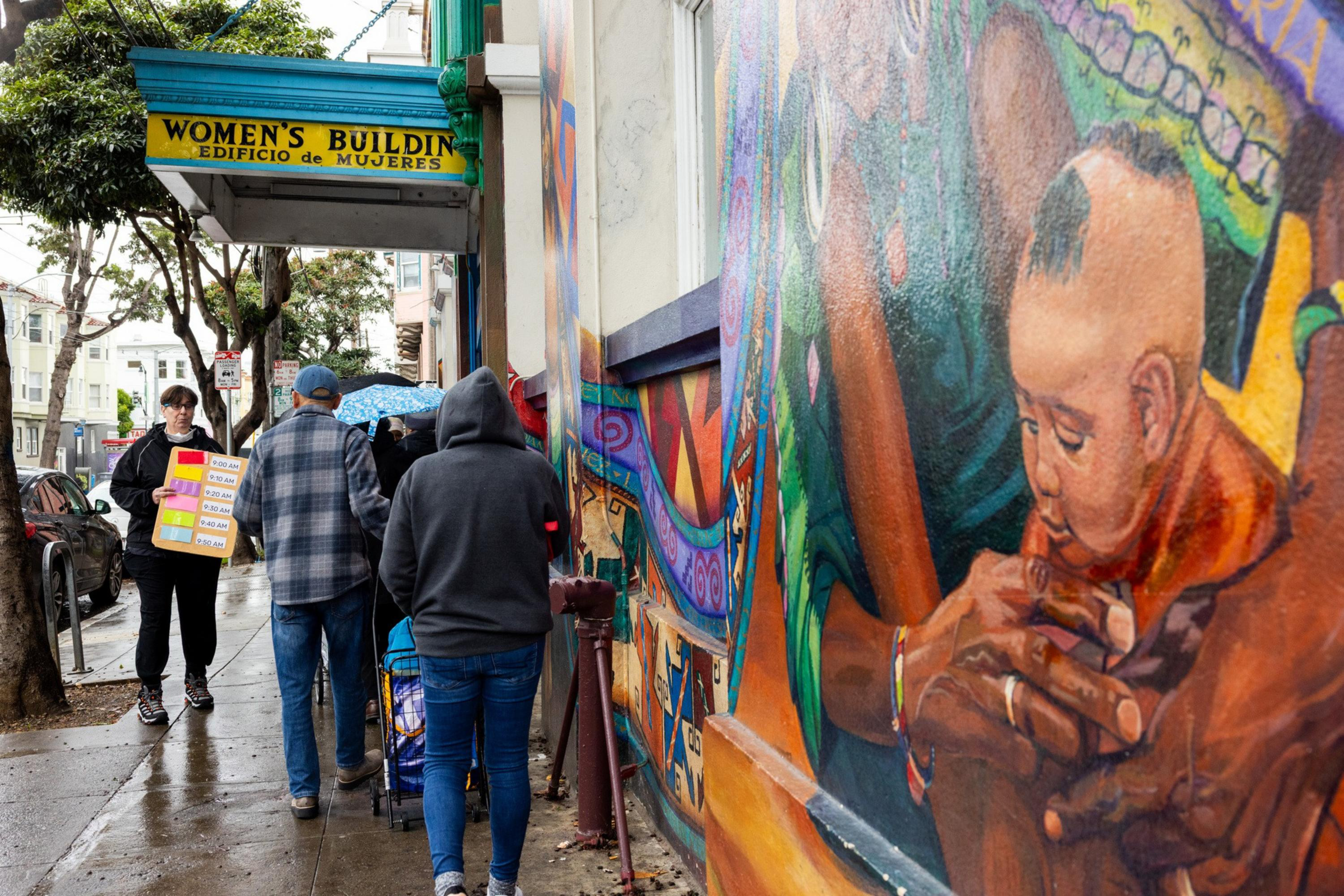 A group of people walk past a colorful mural of a mother holding a baby, near a building with a sign reading "Women's Building / Edificio de Mujeres."