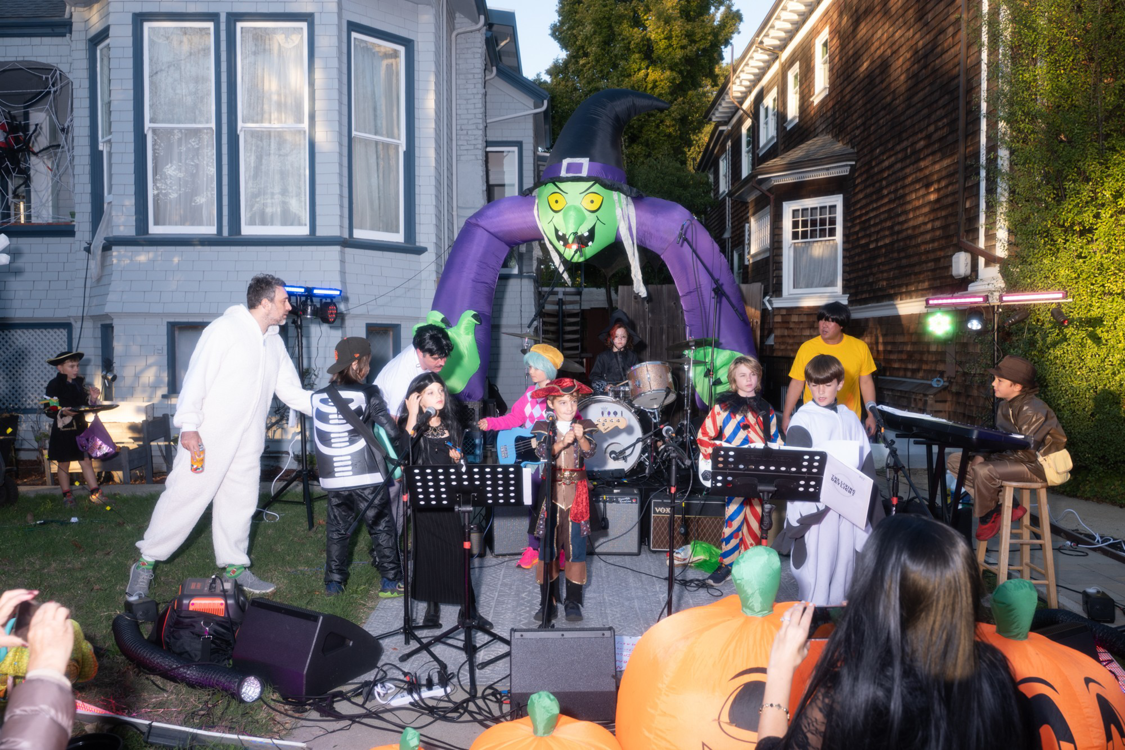 Children and adults in Halloween costumes perform music on an outdoor stage decorated with an inflatable witch and large pumpkin props.