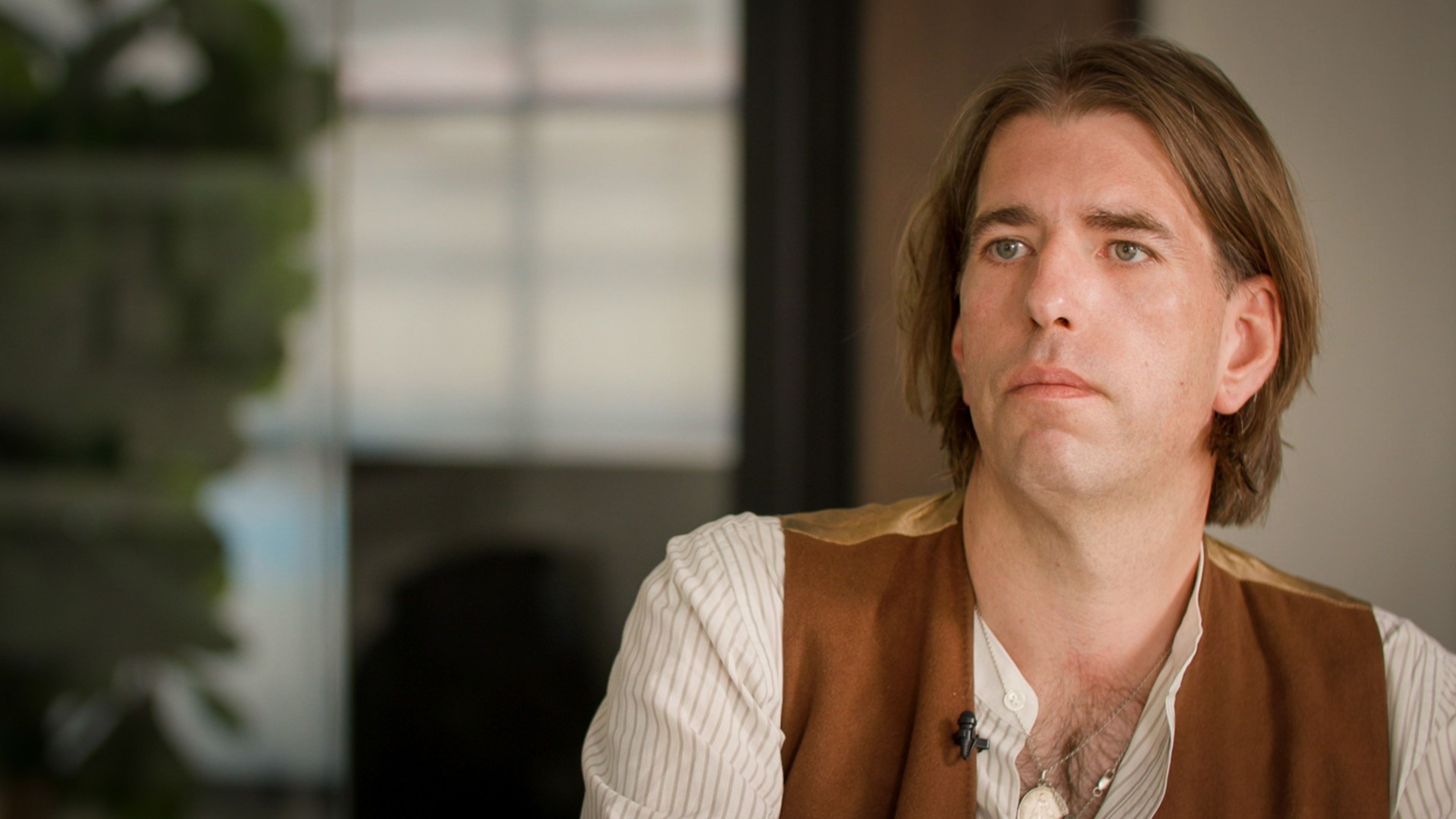 A man with shoulder-length brown hair wears a white striped shirt and brown vest, looking thoughtfully to the side in a softly lit indoor setting.