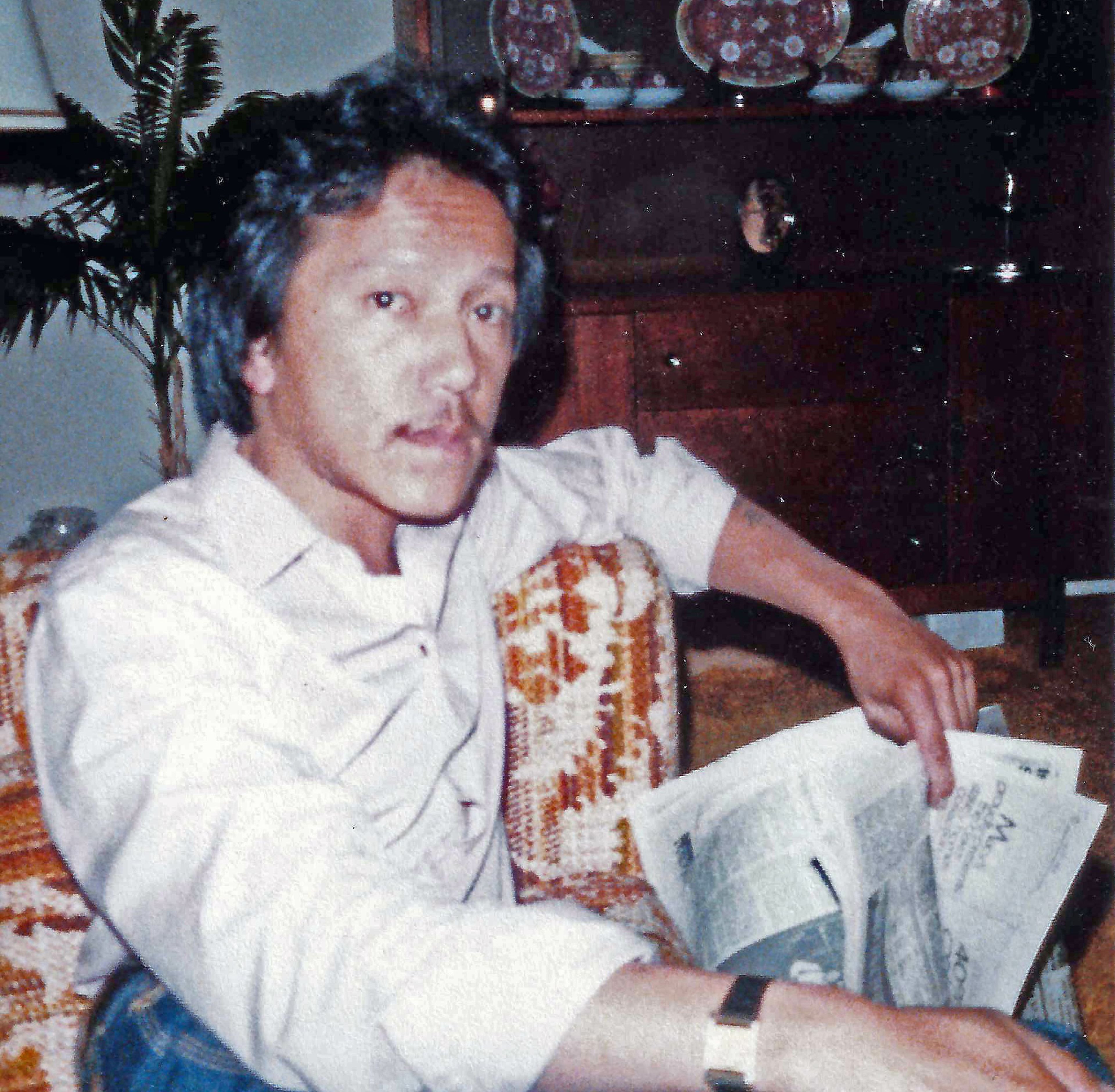 A man with dark hair wearing a light shirt sits on a patterned couch, holding an open newspaper, with a wooden cabinet and plants in the background.