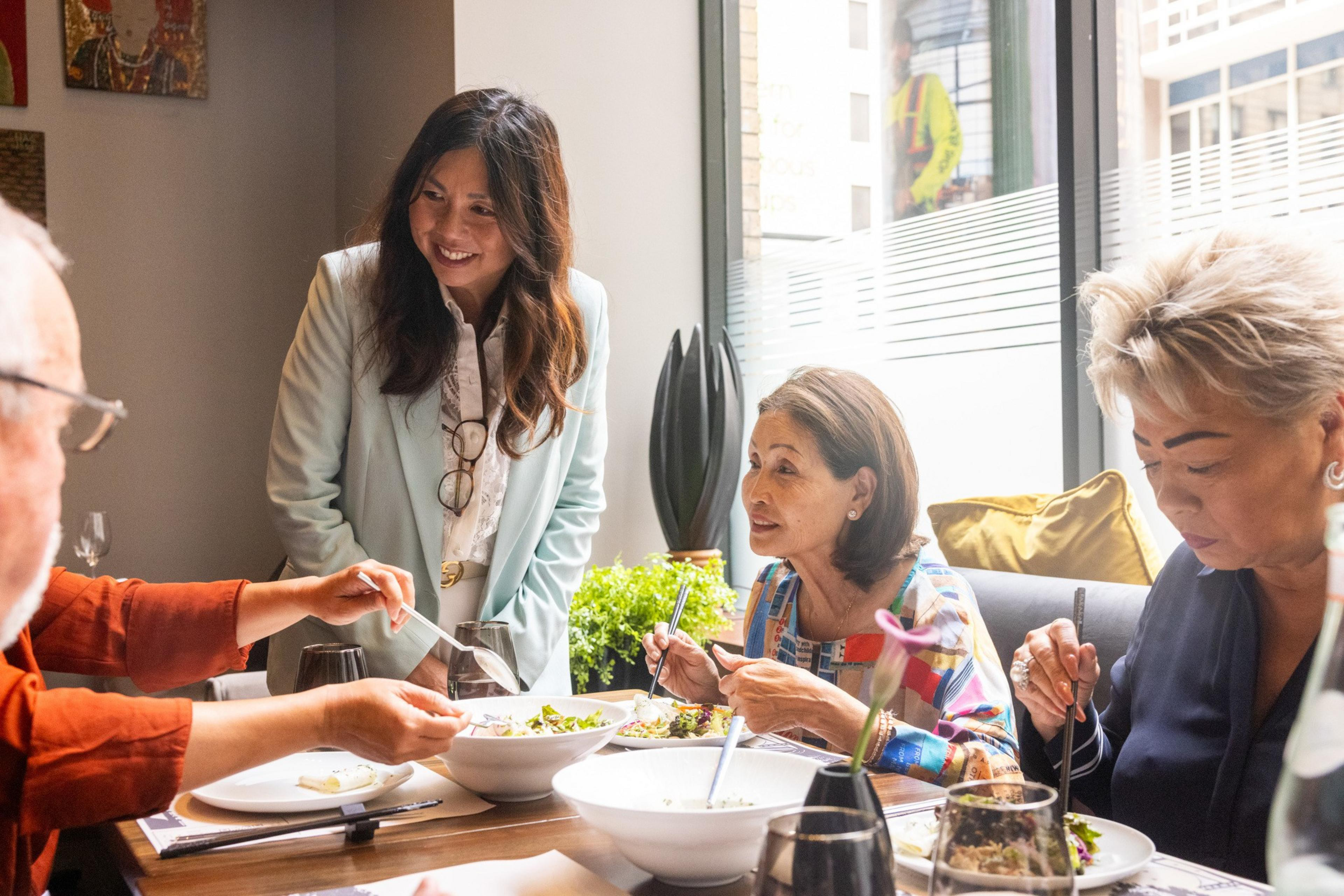 Four people are gathered at a table in a bright room, sharing food and engaging in conversation, with one woman standing and smiling.