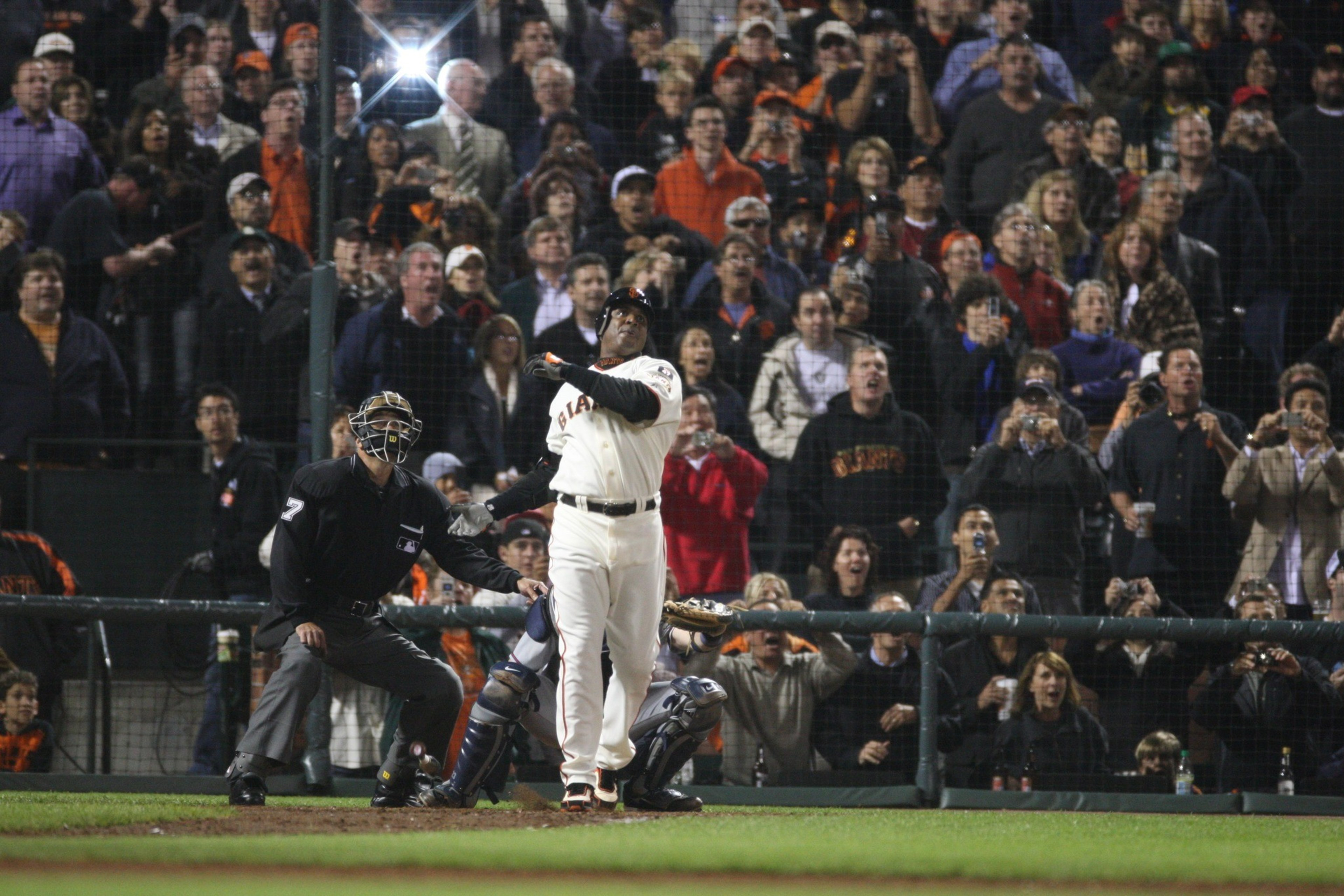 A baseball player in a white Giants uniform just hit the ball as the umpire and catcher watch, with a crowd of spectators reacting behind them.