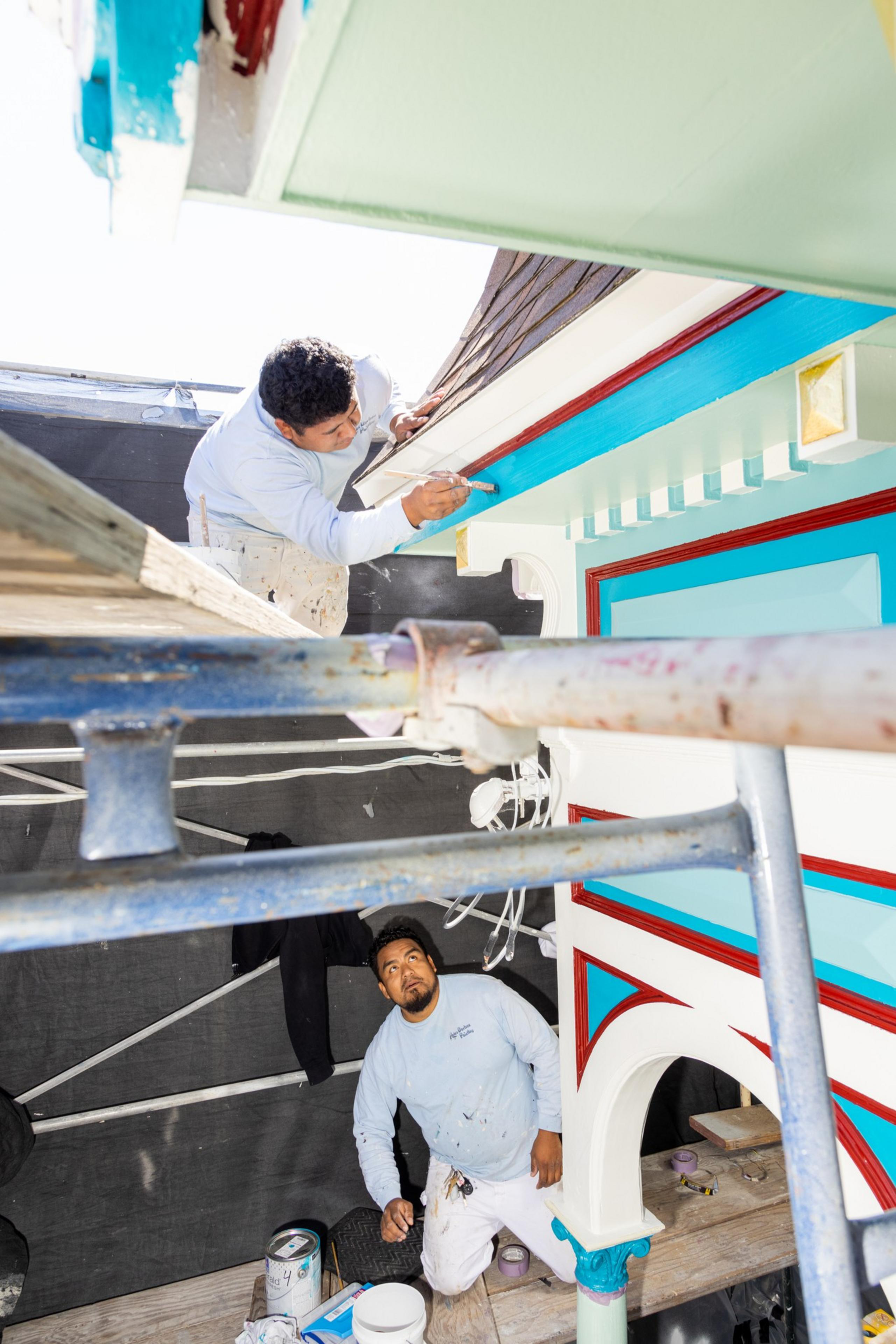 Two painters, one on a ladder and one below, are working on colorful architectural details.