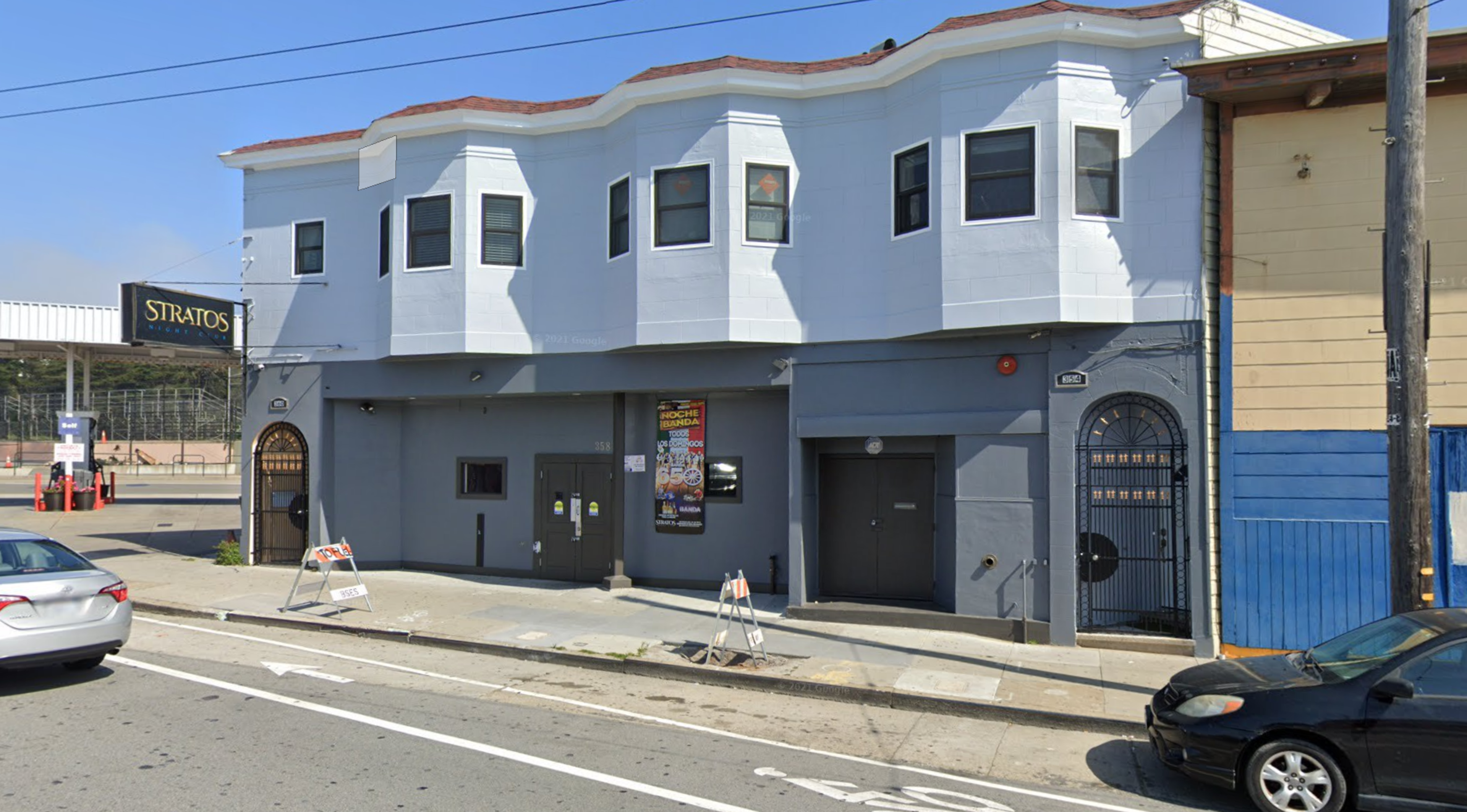 A two-story building with a white upper level and gray lower level has a sign reading "STRATOS" on the left and parked cars on either side of the street.