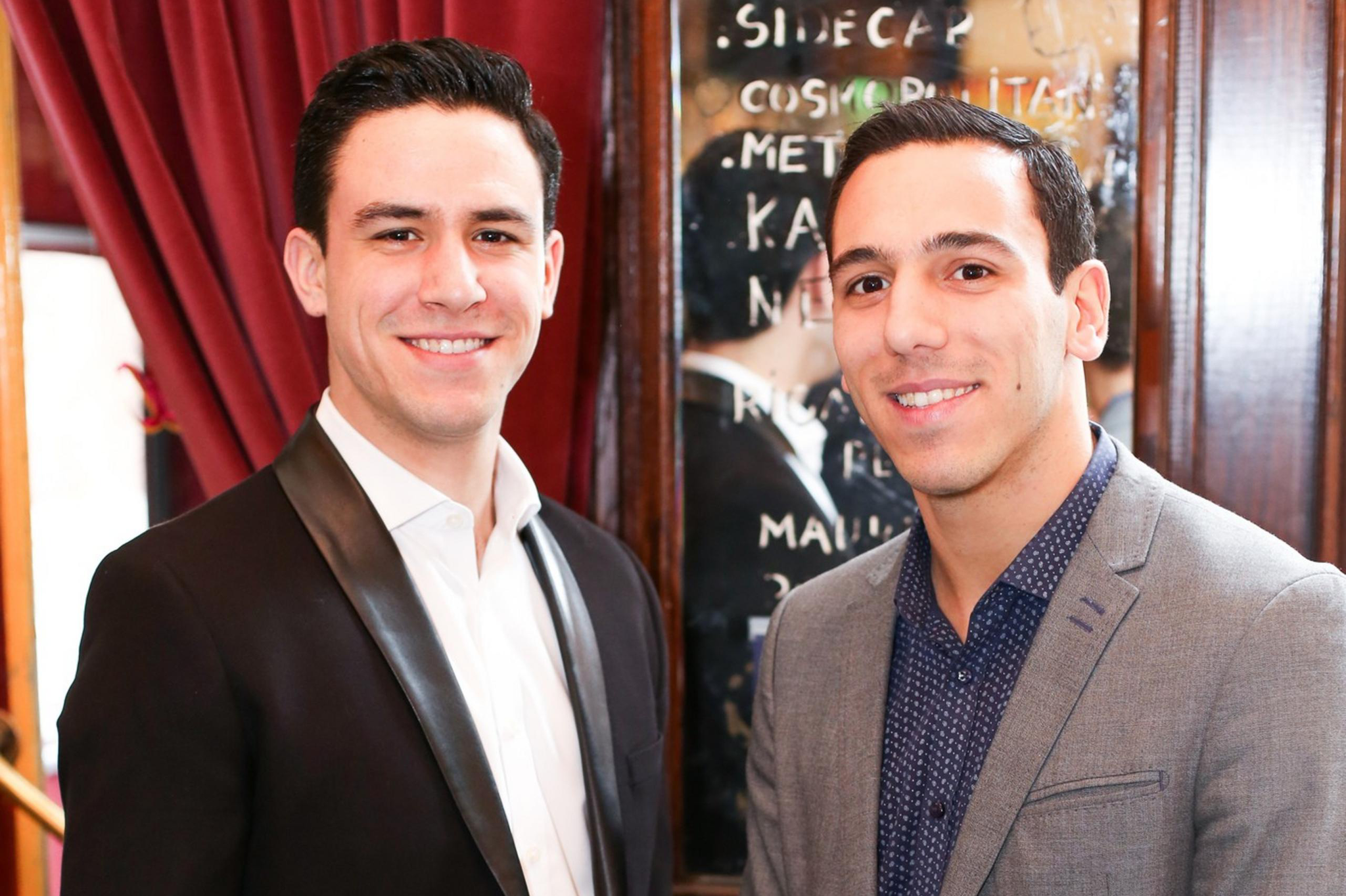 Two young men stand side by side indoors, smiling at the camera, dressed in blazers with a wooden wall and a reflective menu behind them.