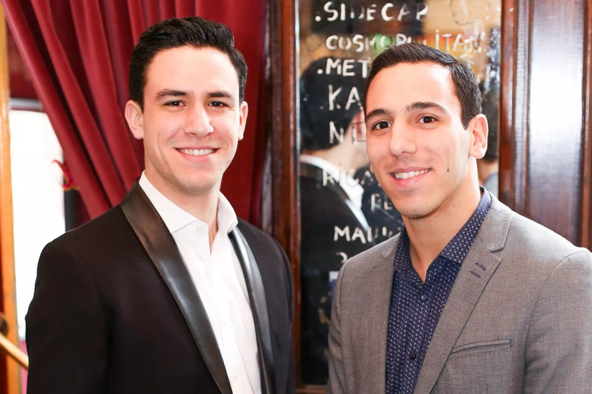 Two young men stand side by side indoors, smiling at the camera, dressed in blazers with a wooden wall and a reflective menu behind them.