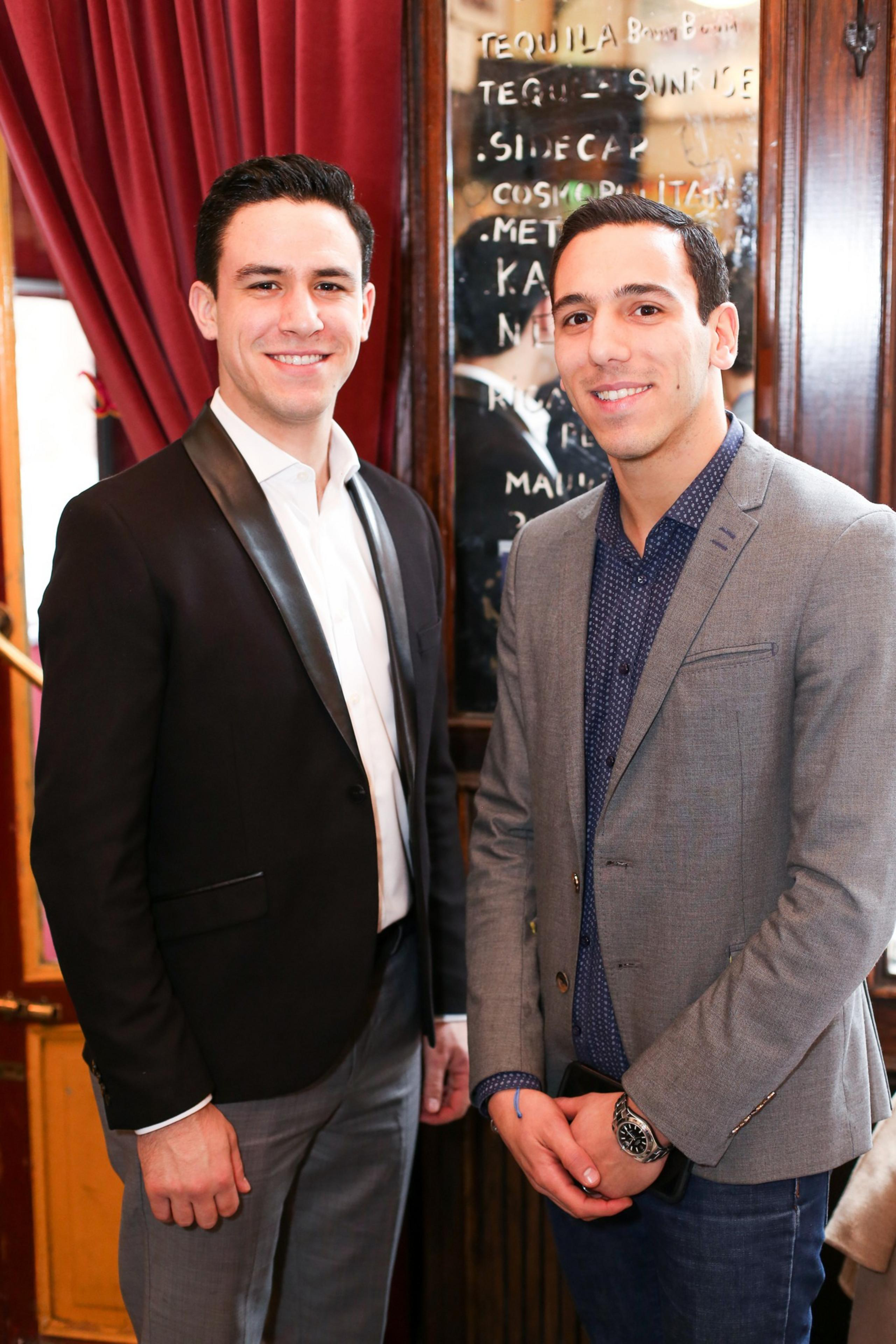 Two young men stand side by side indoors, smiling at the camera, dressed in blazers with a wooden wall and a reflective menu behind them.
