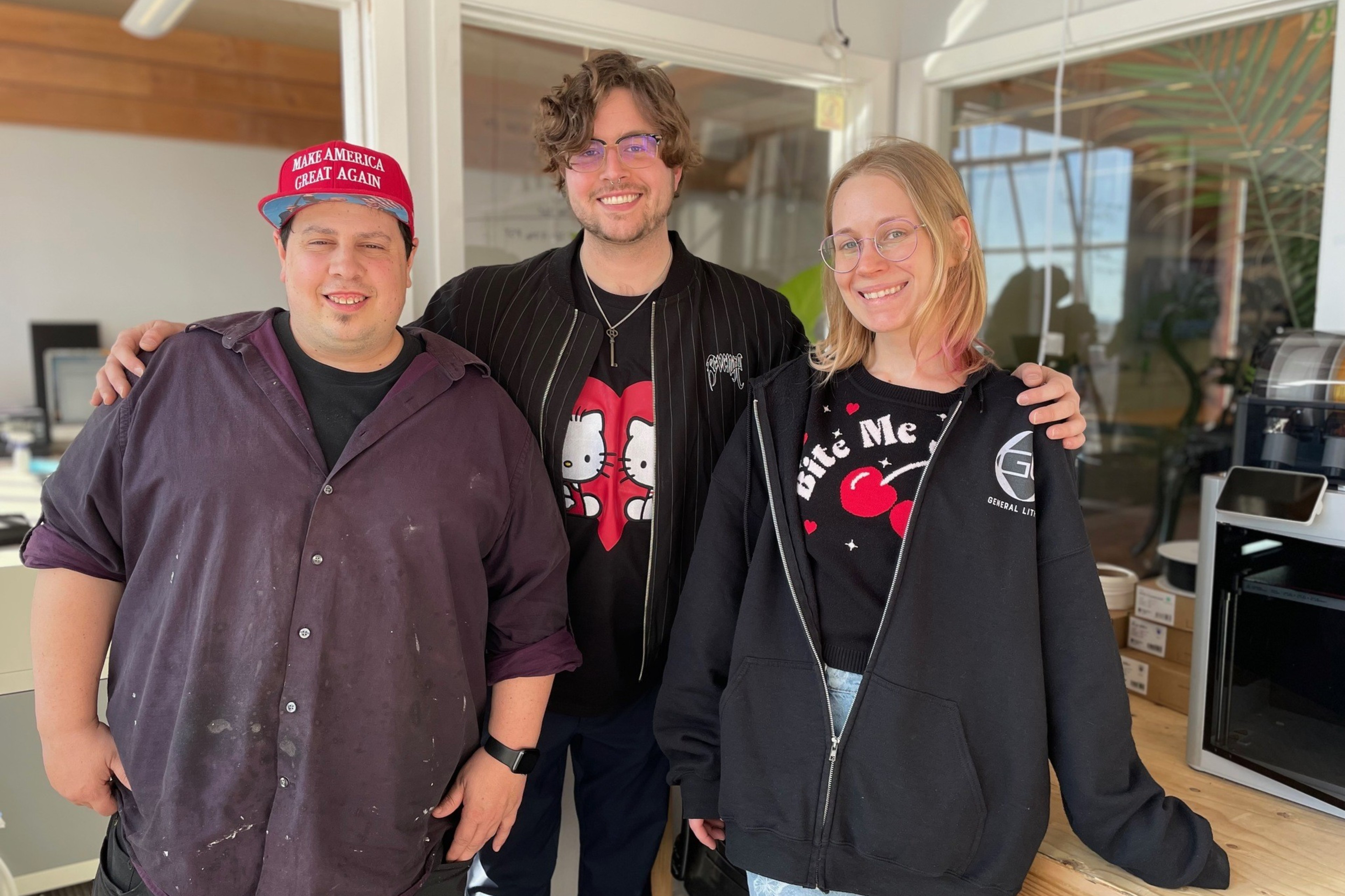 Three people are smiling in an office. One wears a red hat, another has a cartoon shirt, and the third is wearing glasses and a hoodie.