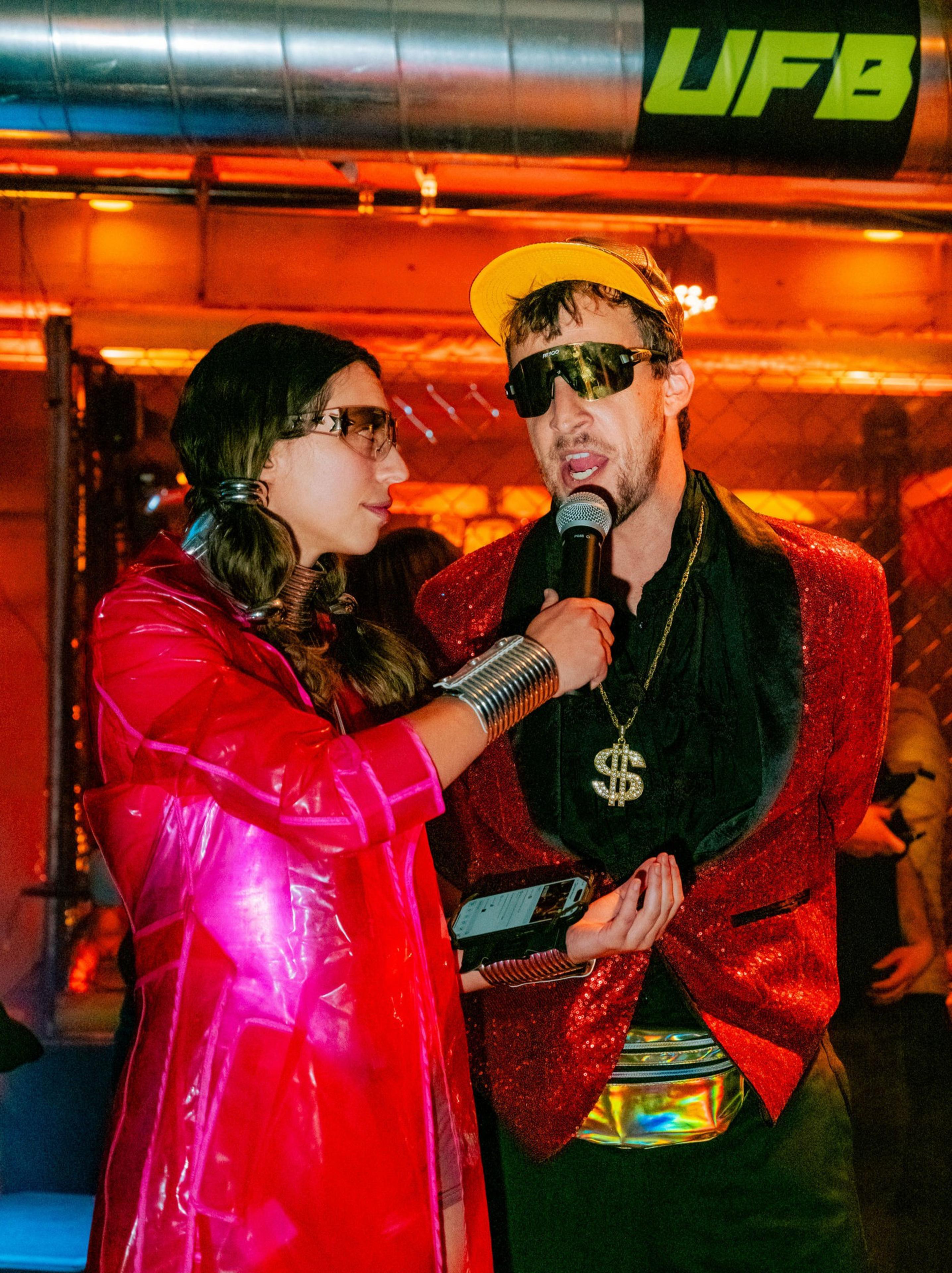 A woman in a shiny pink coat holds a microphone to a man wearing a red glittery jacket, dollar sign necklace, yellow cap, and dark sunglasses.