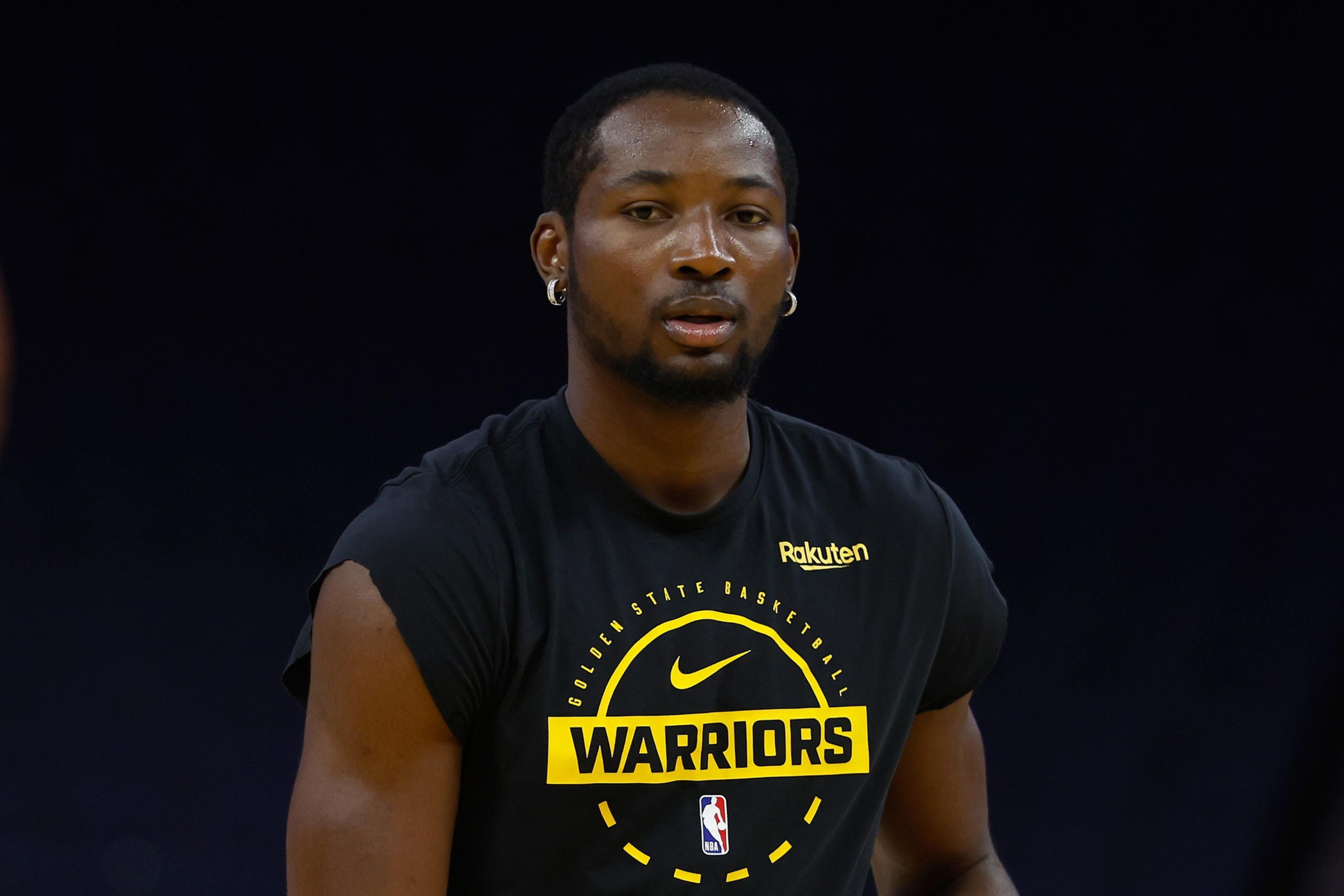 A man wearing a black sleeveless Golden State Warriors shirt looks forward against a dark background.