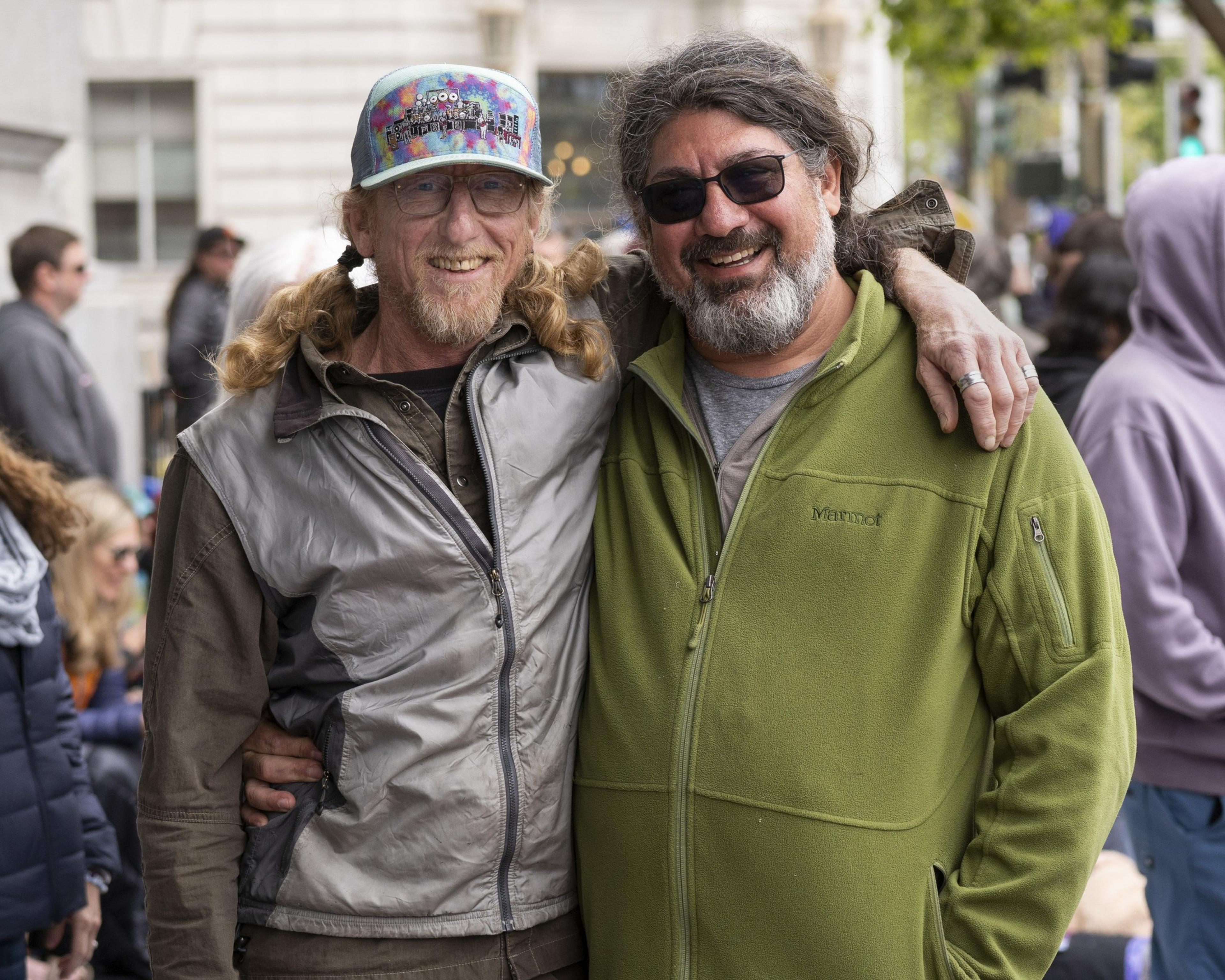 Two men are smiling and embracing. One wears a colorful cap and glasses, the other wears a green jacket. They are outdoors, surrounded by people.